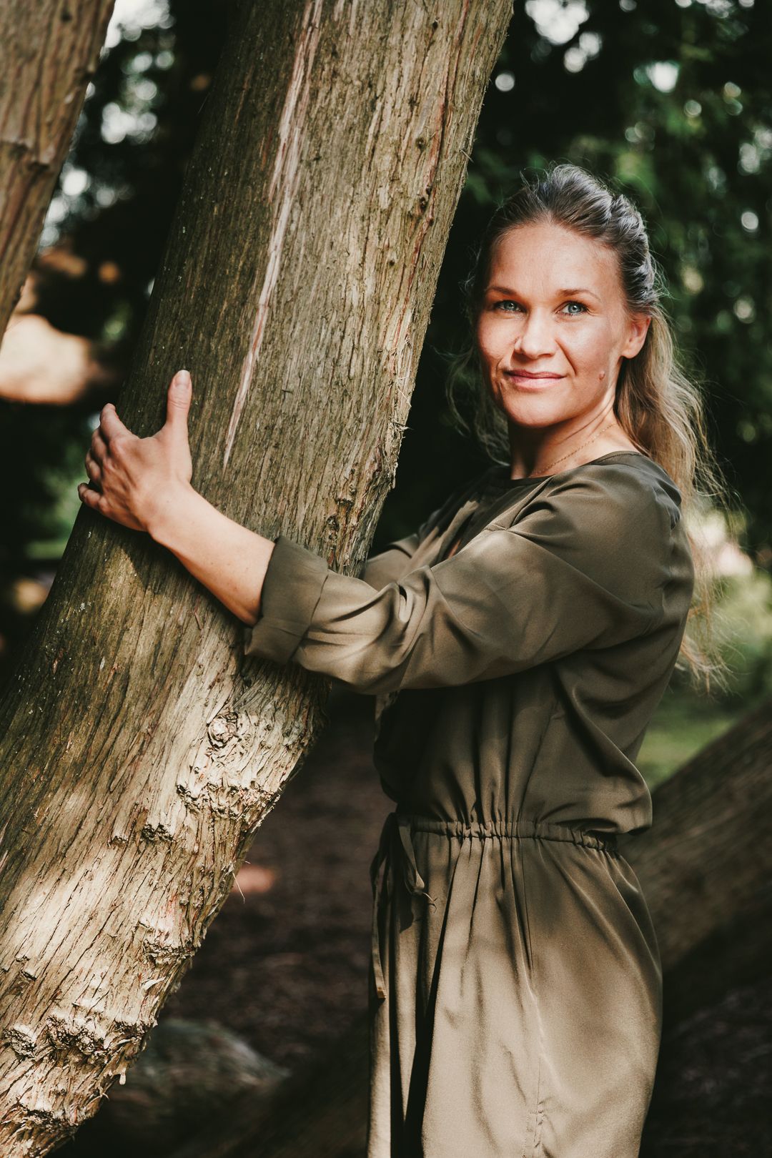 Woman in olive green dress hugging a tree trunk in a lush forest setting.