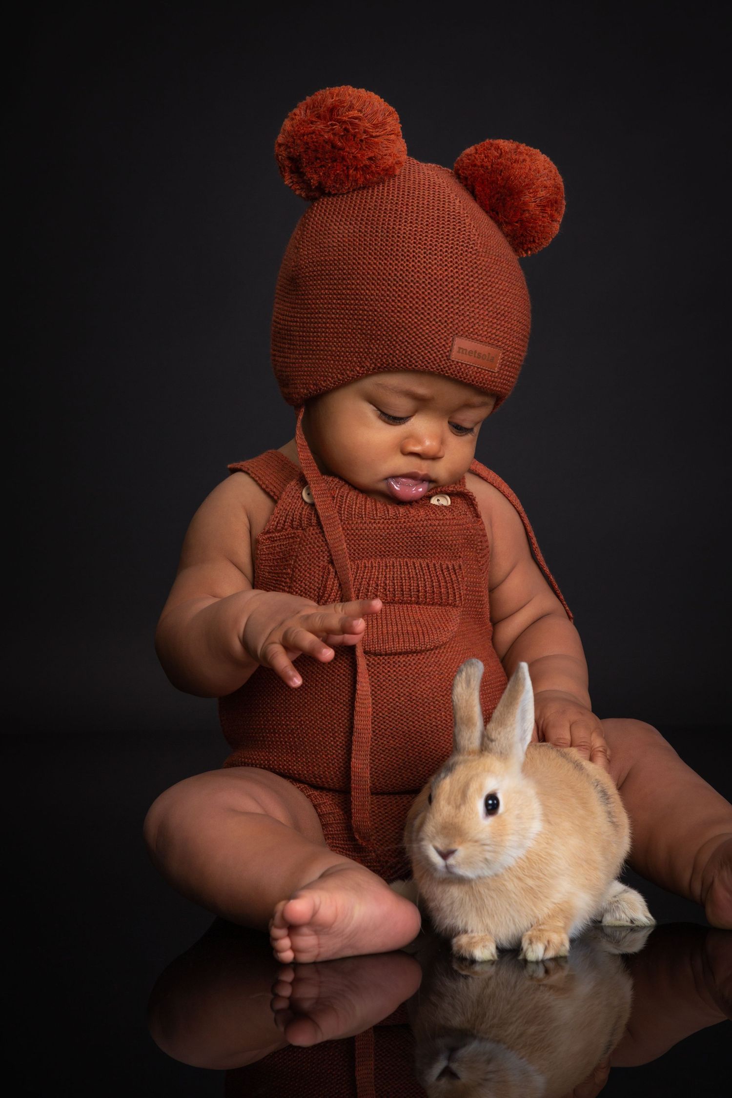 Baby in rust knit bear hat and romper gently touches a small tan rabbit against a dark background.