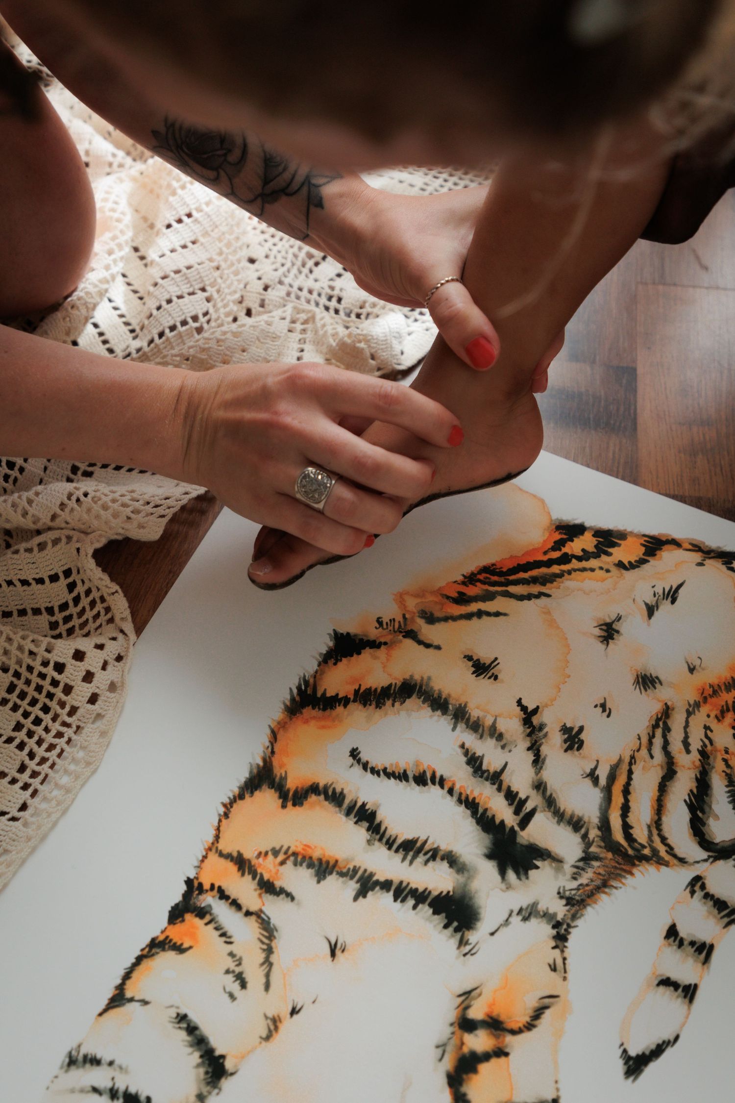 Two hands with rings hold a foot above a vibrant watercolor tiger painting on white paper.