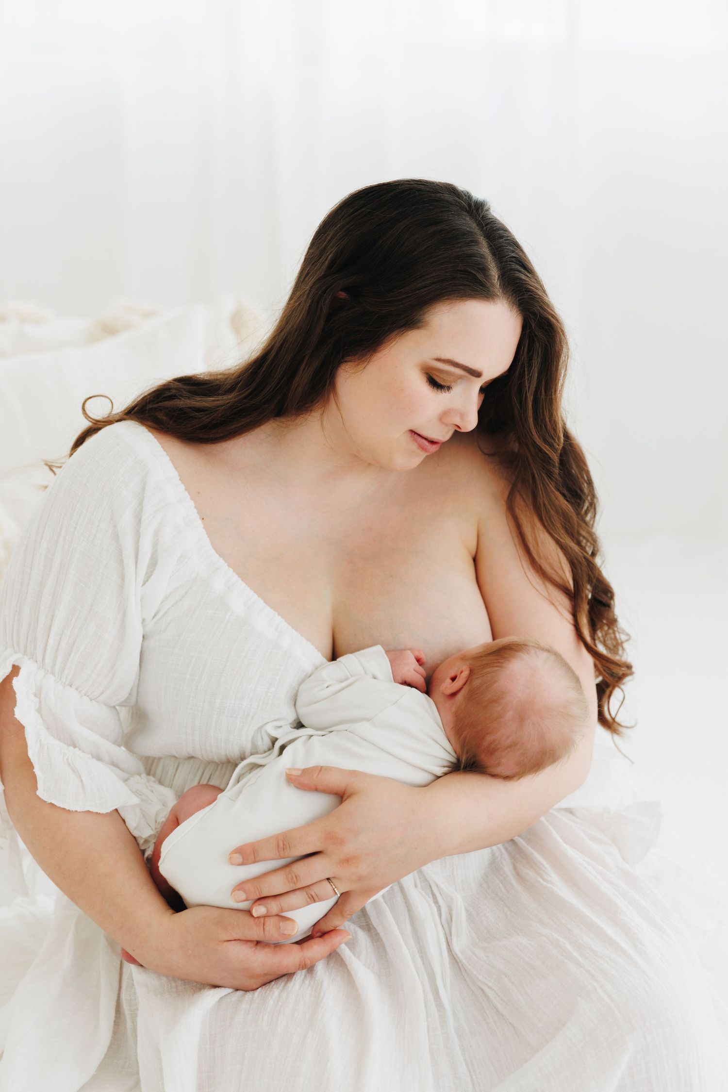 A mother tenderly breastfeeding her newborn baby while wearing a white dress in a bright, serene setting.