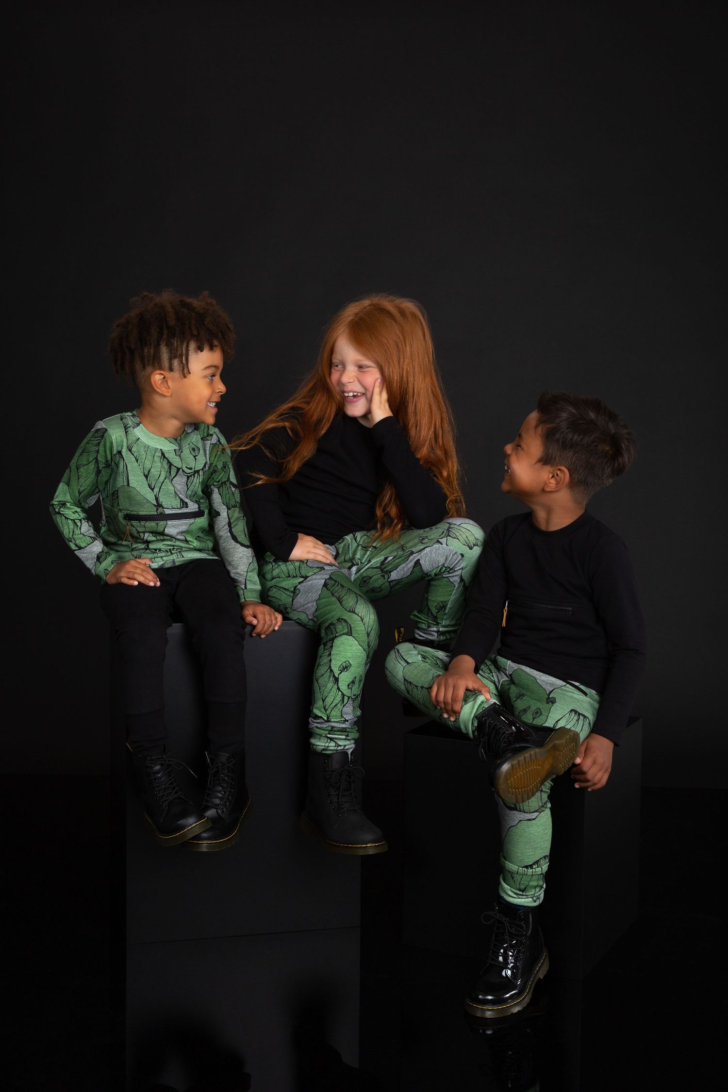 Three children laughing together wearing matching green and black printed tops and leggings against a dark background.