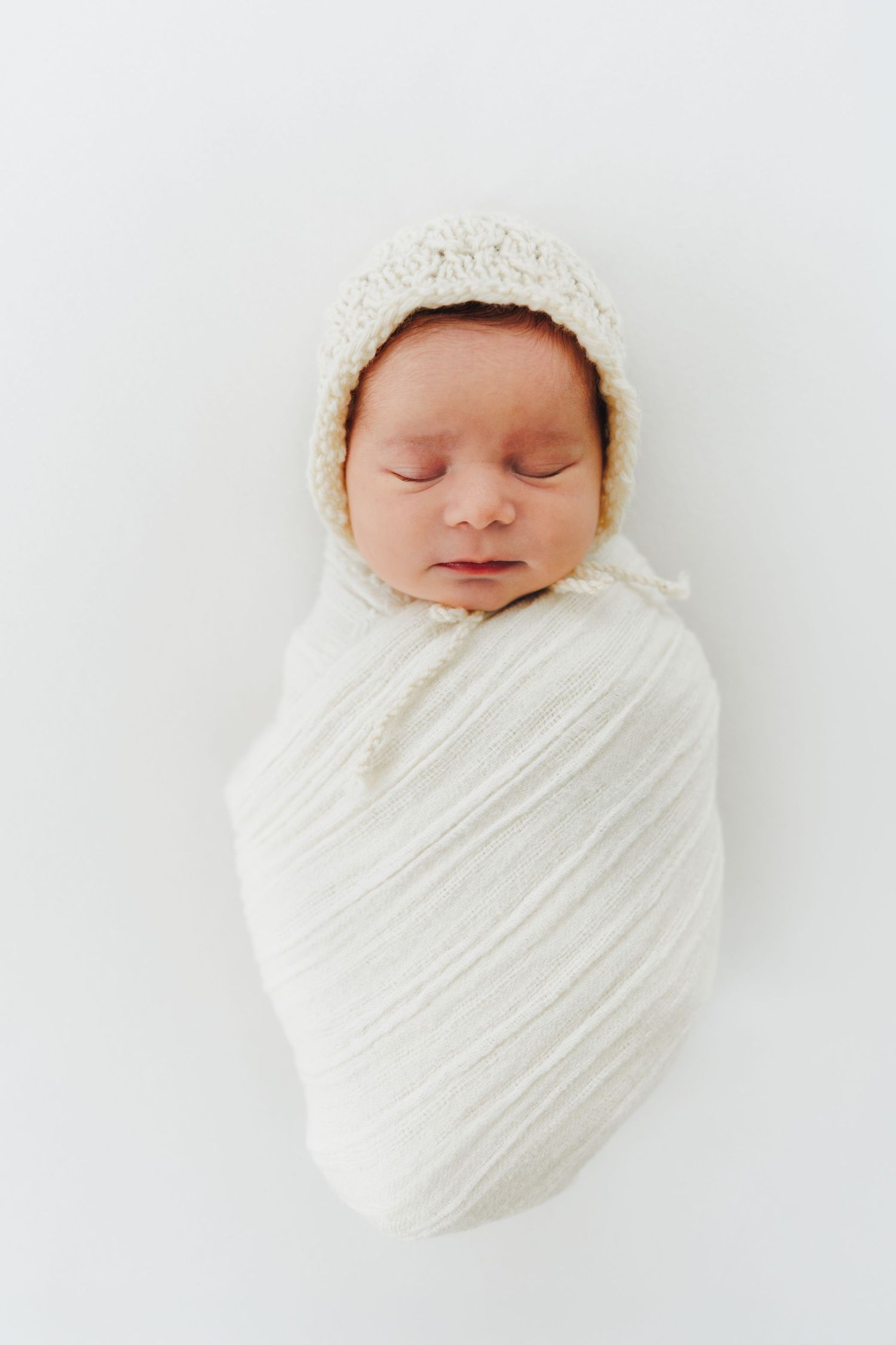 Sleeping newborn baby swaddled in white wrap wearing a cream knit bonnet on a white background. Newborn photography Helsinki