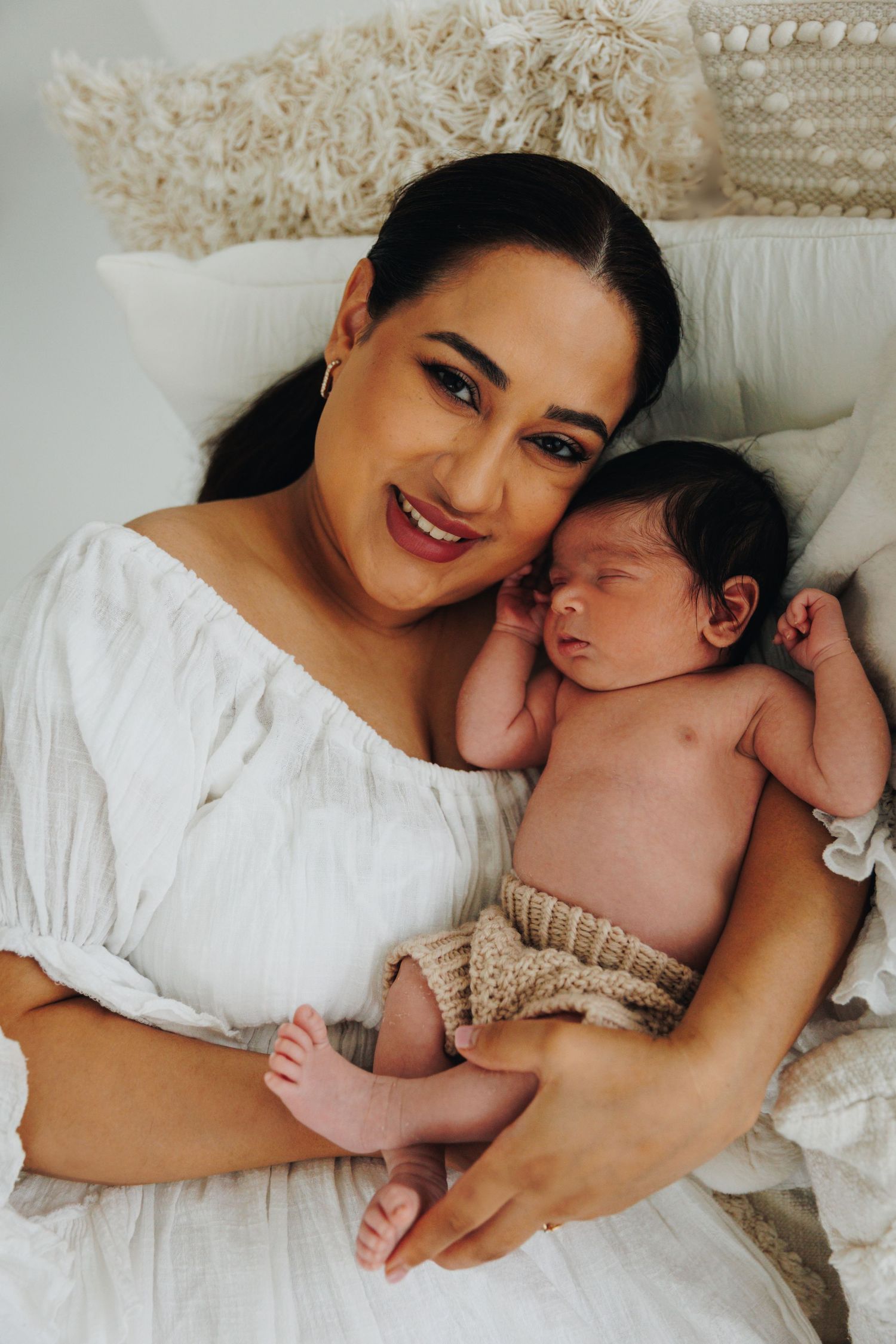A smiling mother in a white dress holds her sleeping newborn baby, posed against soft boho-style cushions.