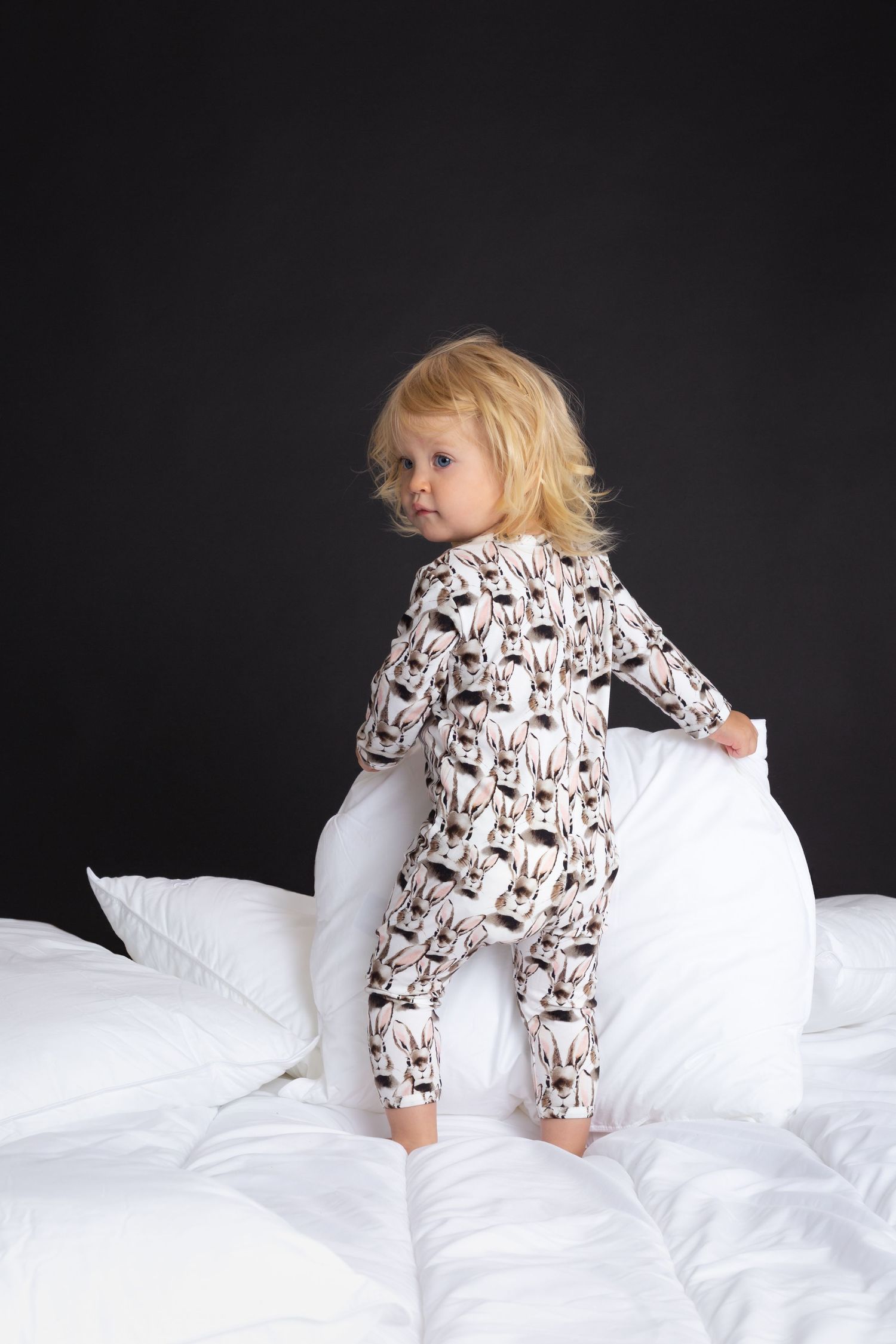 Toddler in patterned pajamas standing on white bedding against dark background.