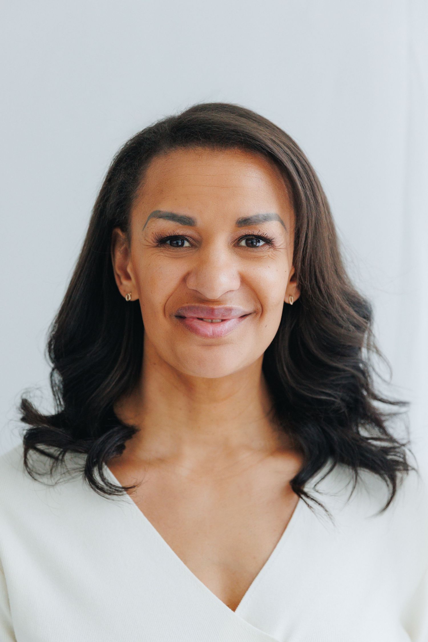 Smiling woman with dark wavy hair wearing a white v-neck top against a light gray background.