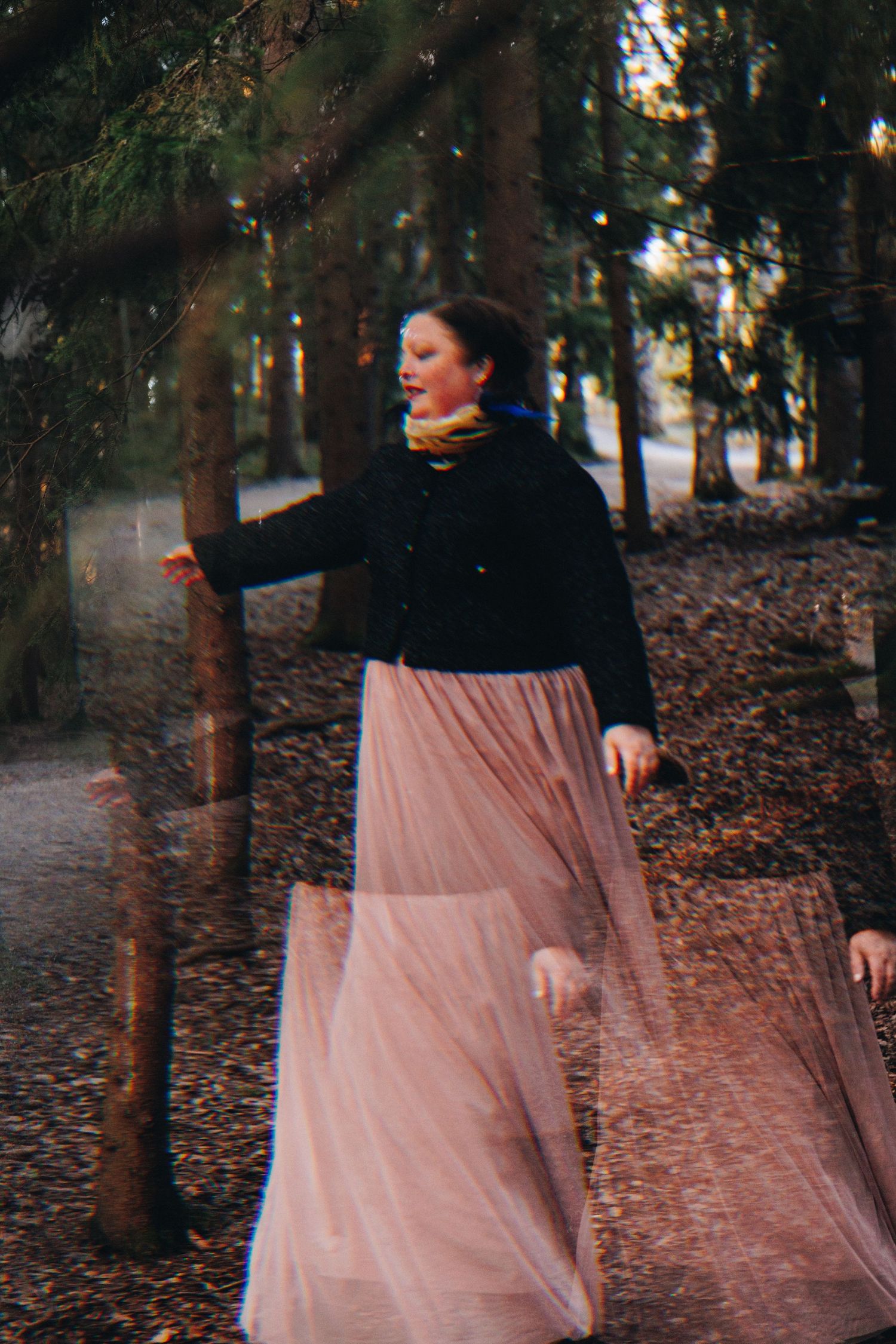 Woman in pink tulle skirt and black jacket dancing gracefully among trees in a forest setting.