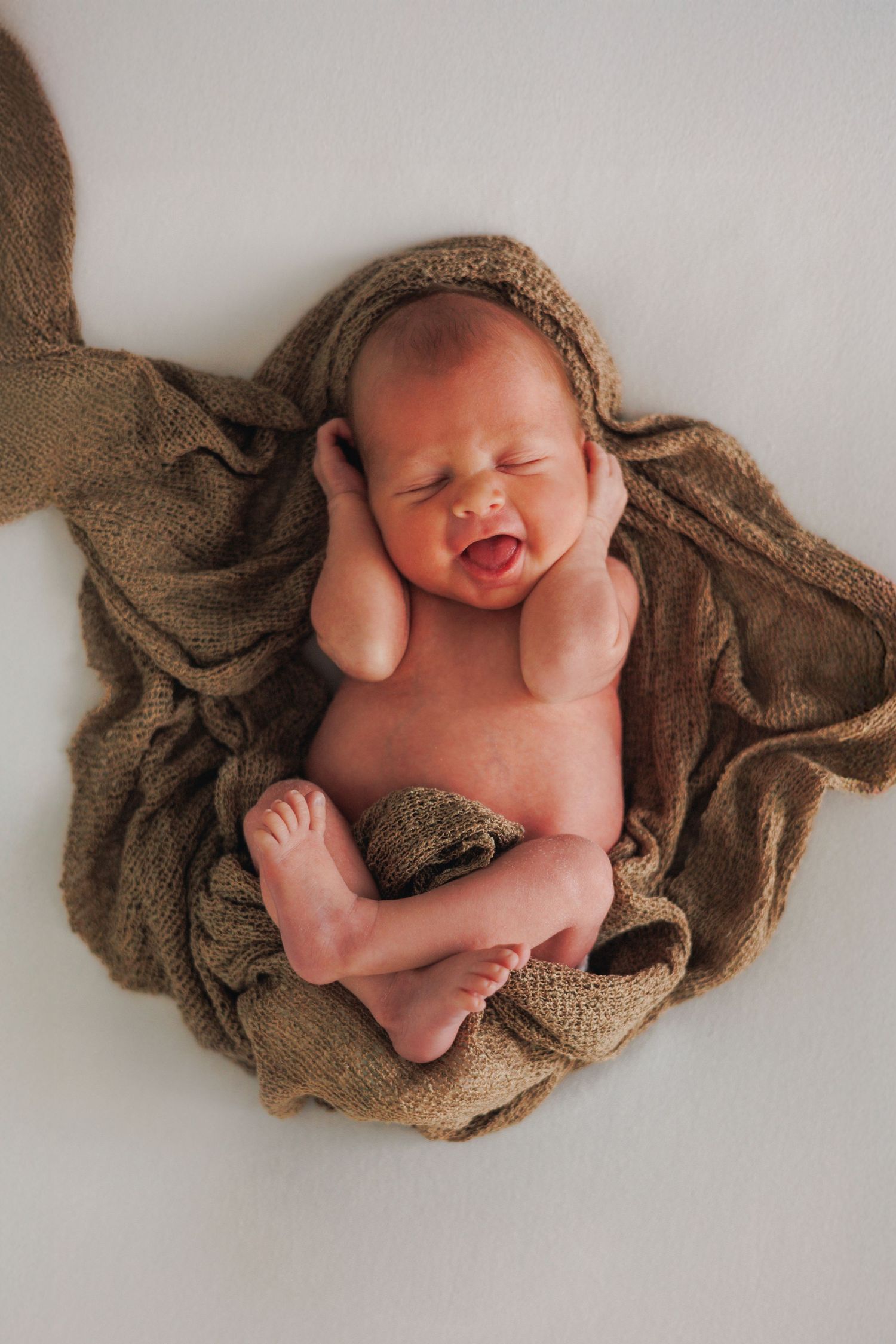 Newborn baby yawning while lying on a brown wrap blanket against a white background.