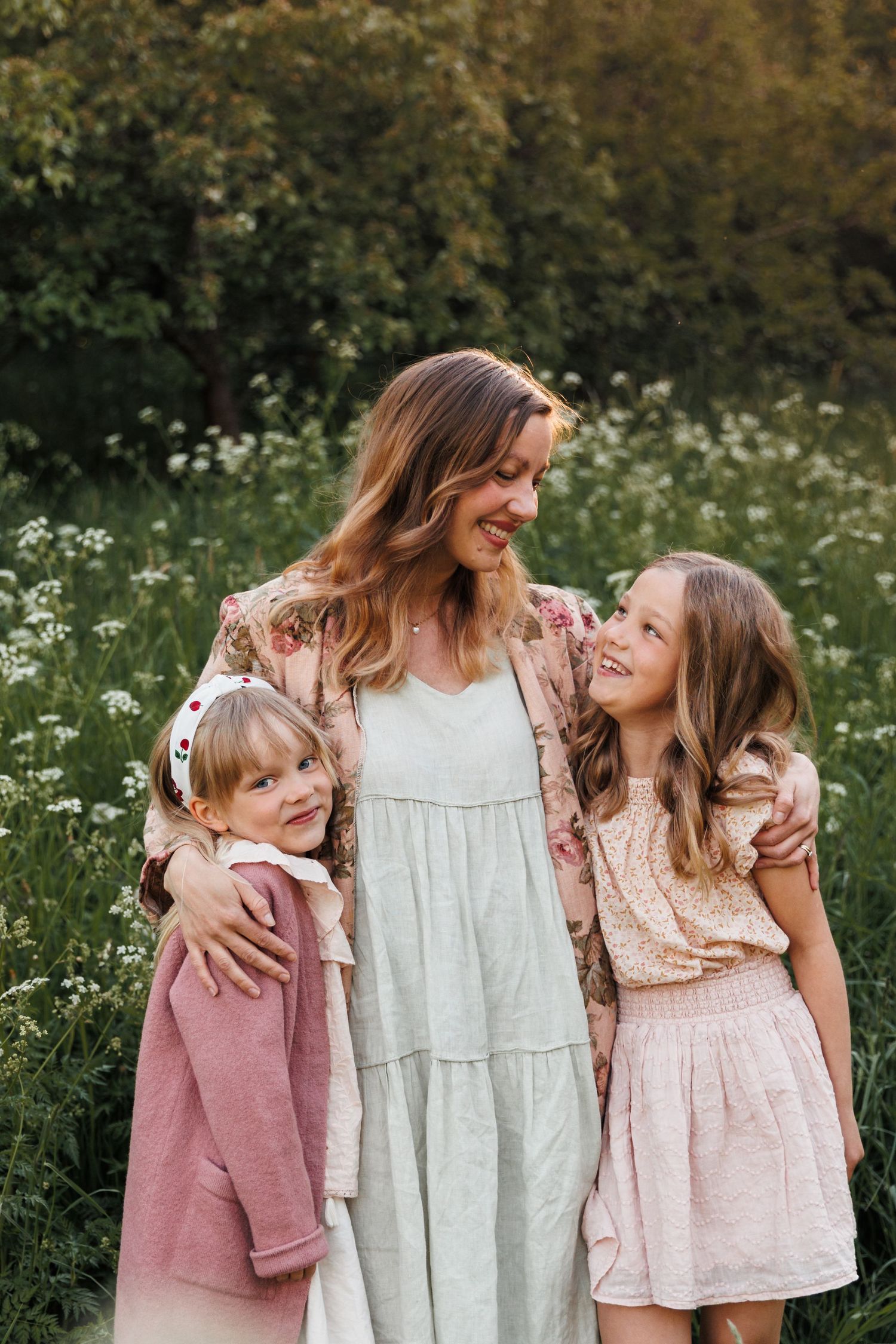 A smiling mother hugs her two young daughters in a sunny wildflower field, all wearing soft pink and floral outfits.