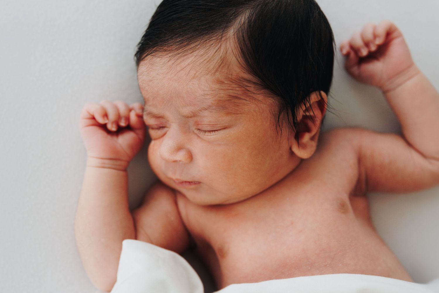 Peaceful newborn baby sleeping with arms raised, dark hair, on a white blanket in a serene setting.