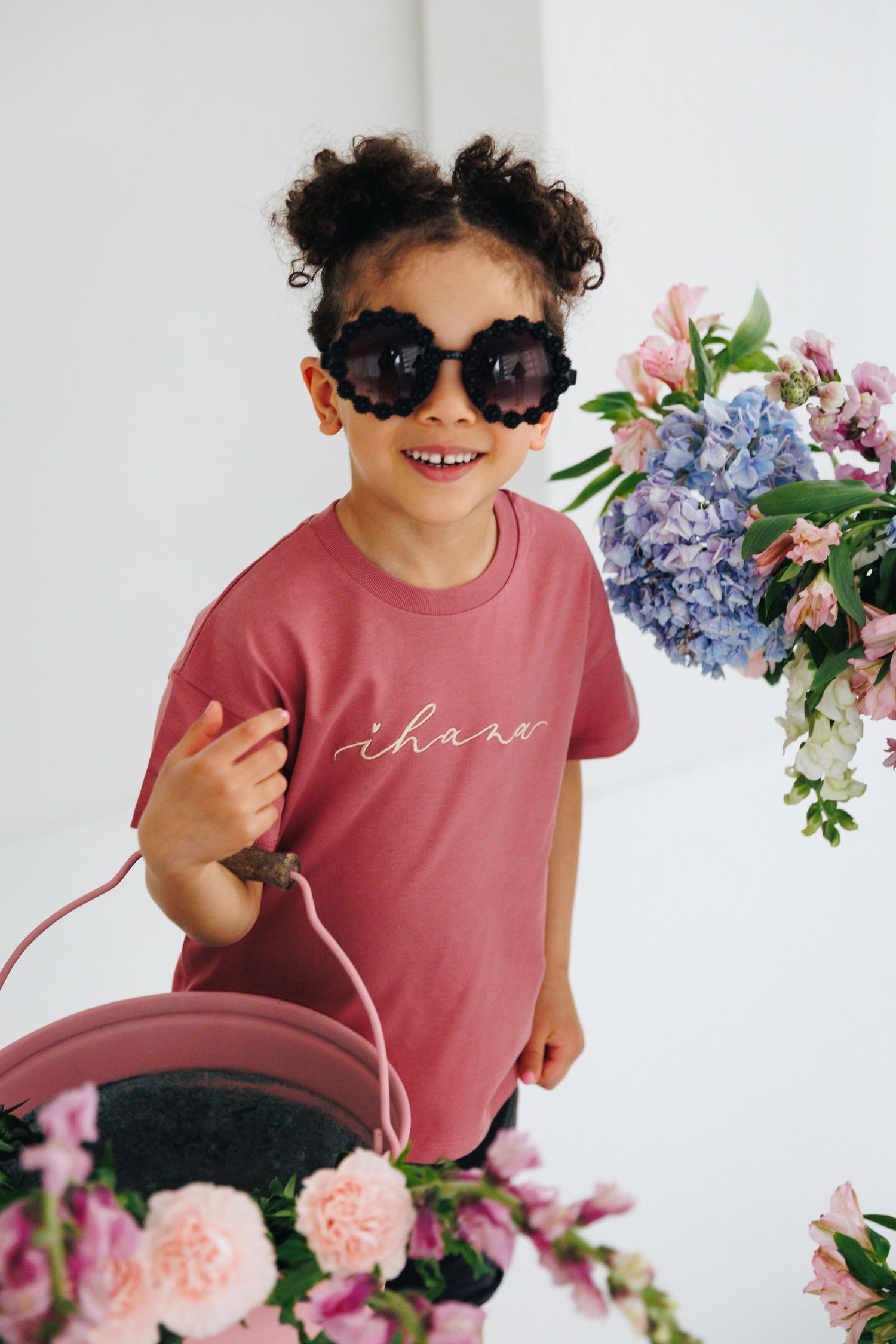 Smiling young girl wearing flower sunglasses and pink t-shirt surrounded by colorful floral arrangements.