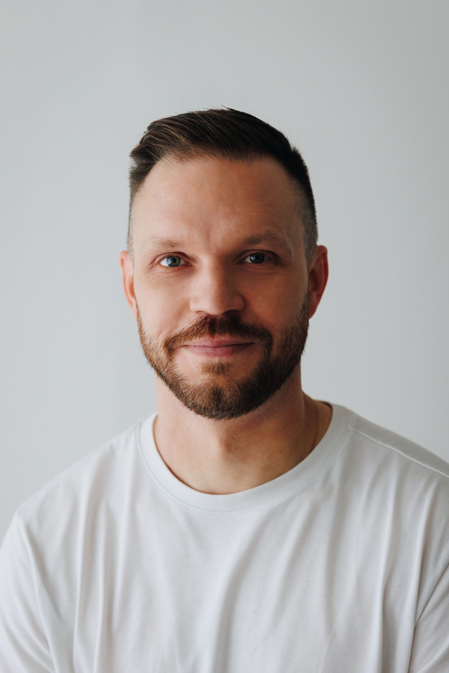 Portrait of person with trimmed beard wearing a white t-shirt against light gray background.