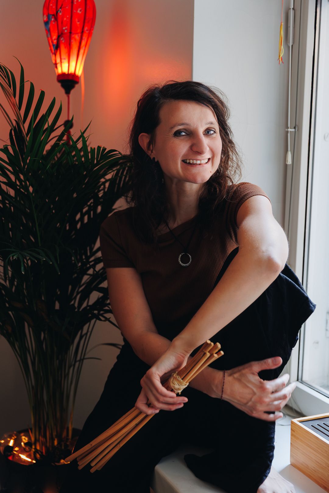 Smiling female massage therapist sitting by a window holding bamboo massage sticks in a cozy spa.