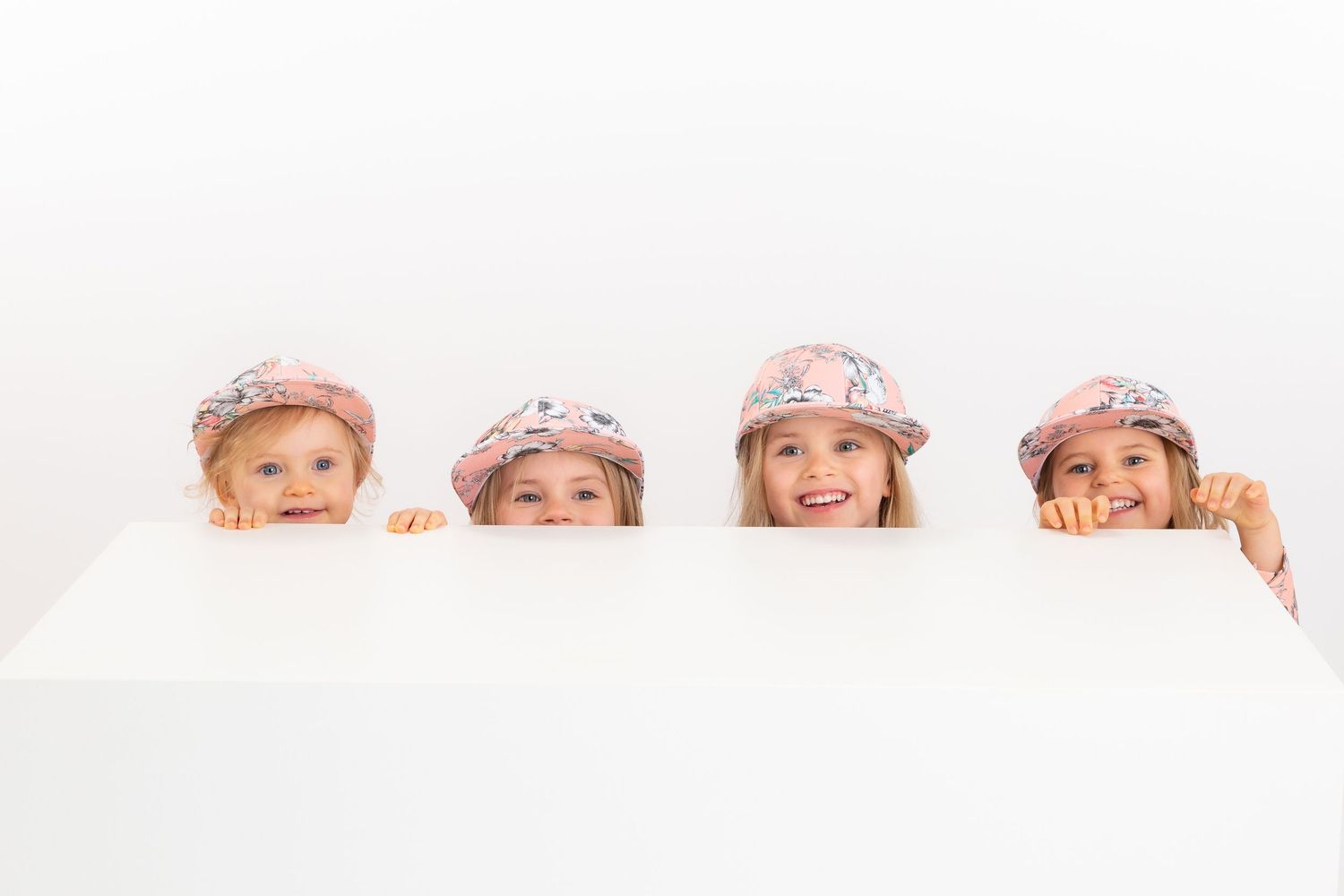 Four young girls wearing matching pink floral caps peek over a white surface, smiling at the camera.
