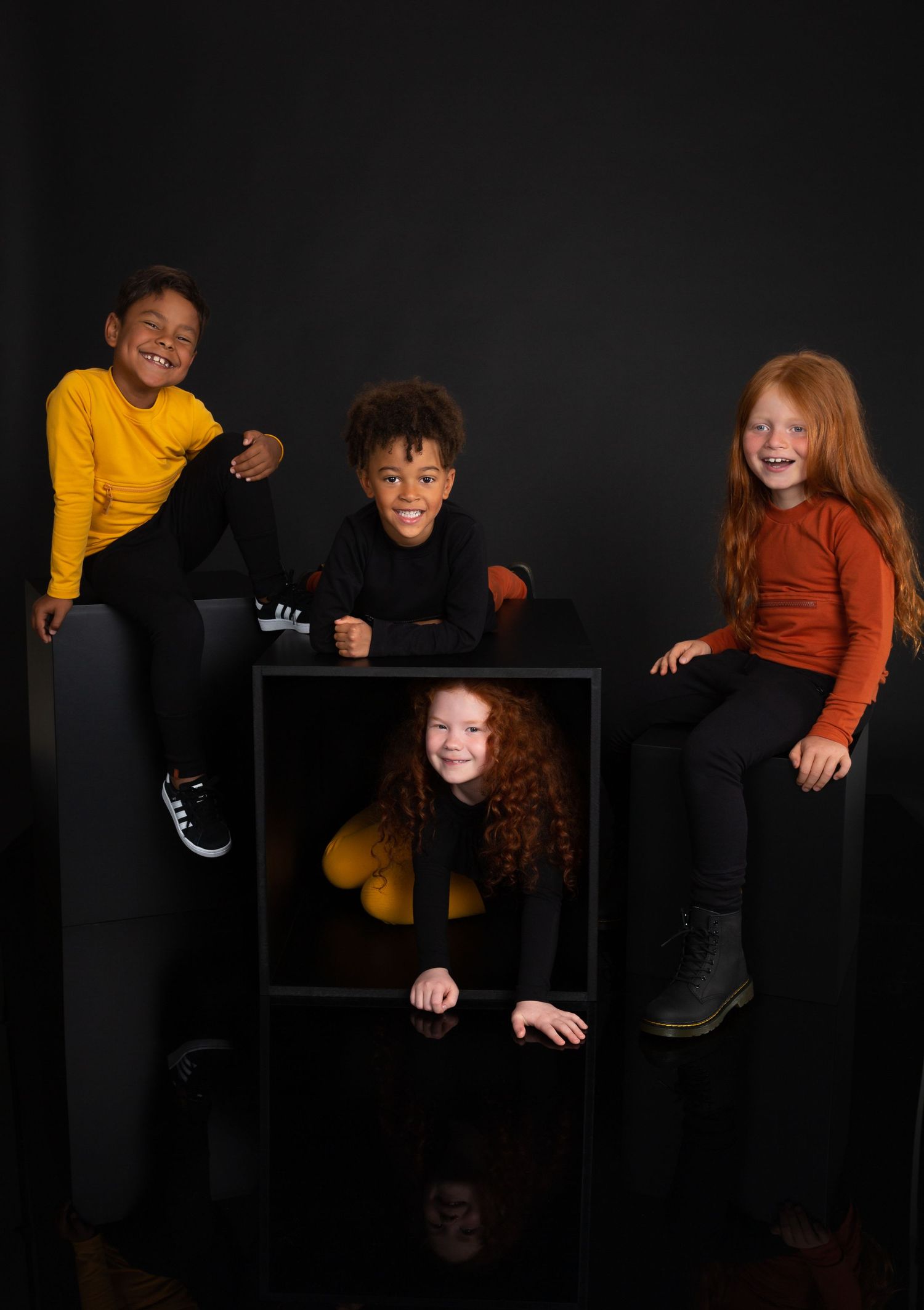 A group of children pose playfully against a black background wearing casual clothing in yellow, brown and black tones.