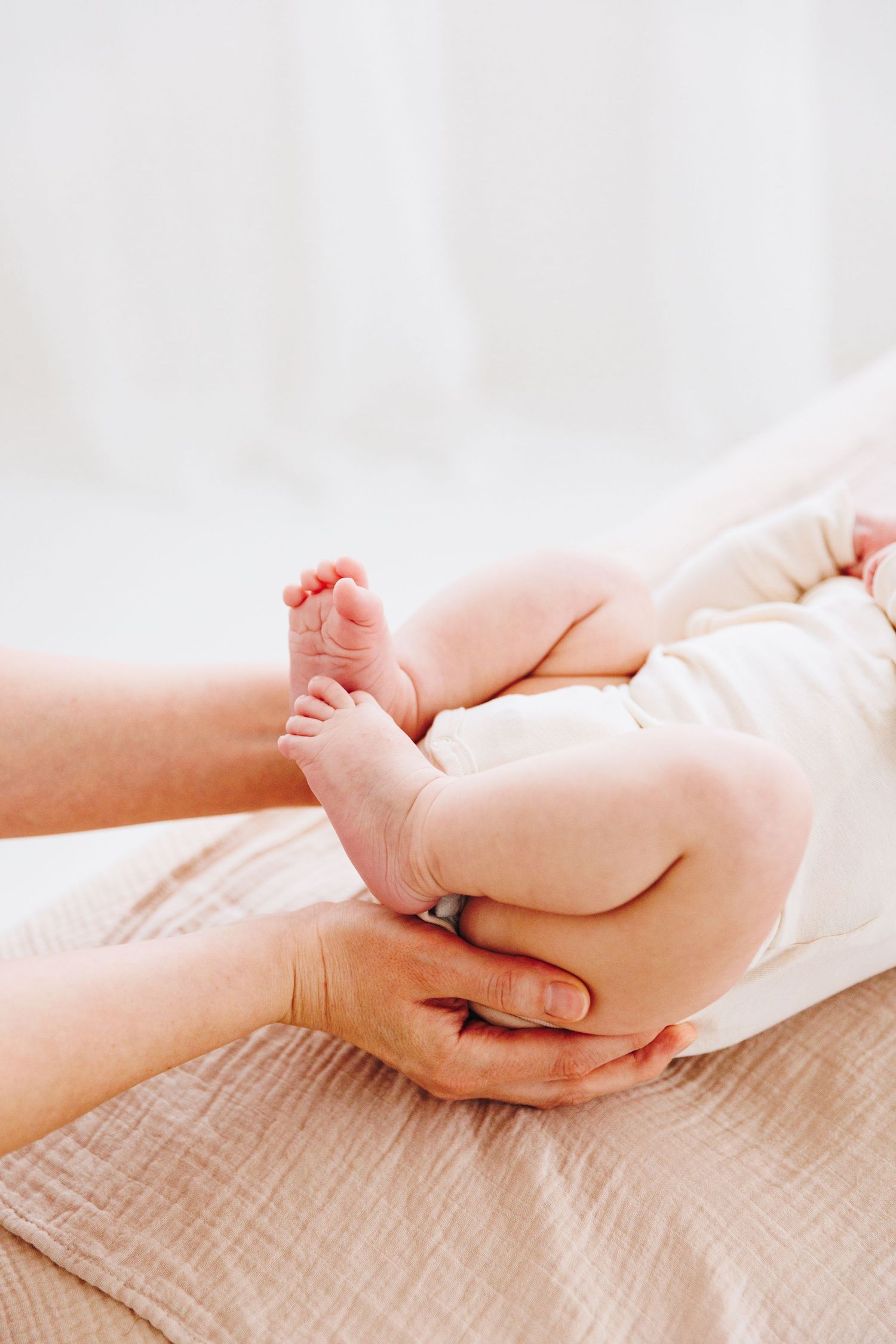 Adult hands gently cradling a newborn baby's tiny feet and bottom, dressed in white onesie on a soft beige blanket.