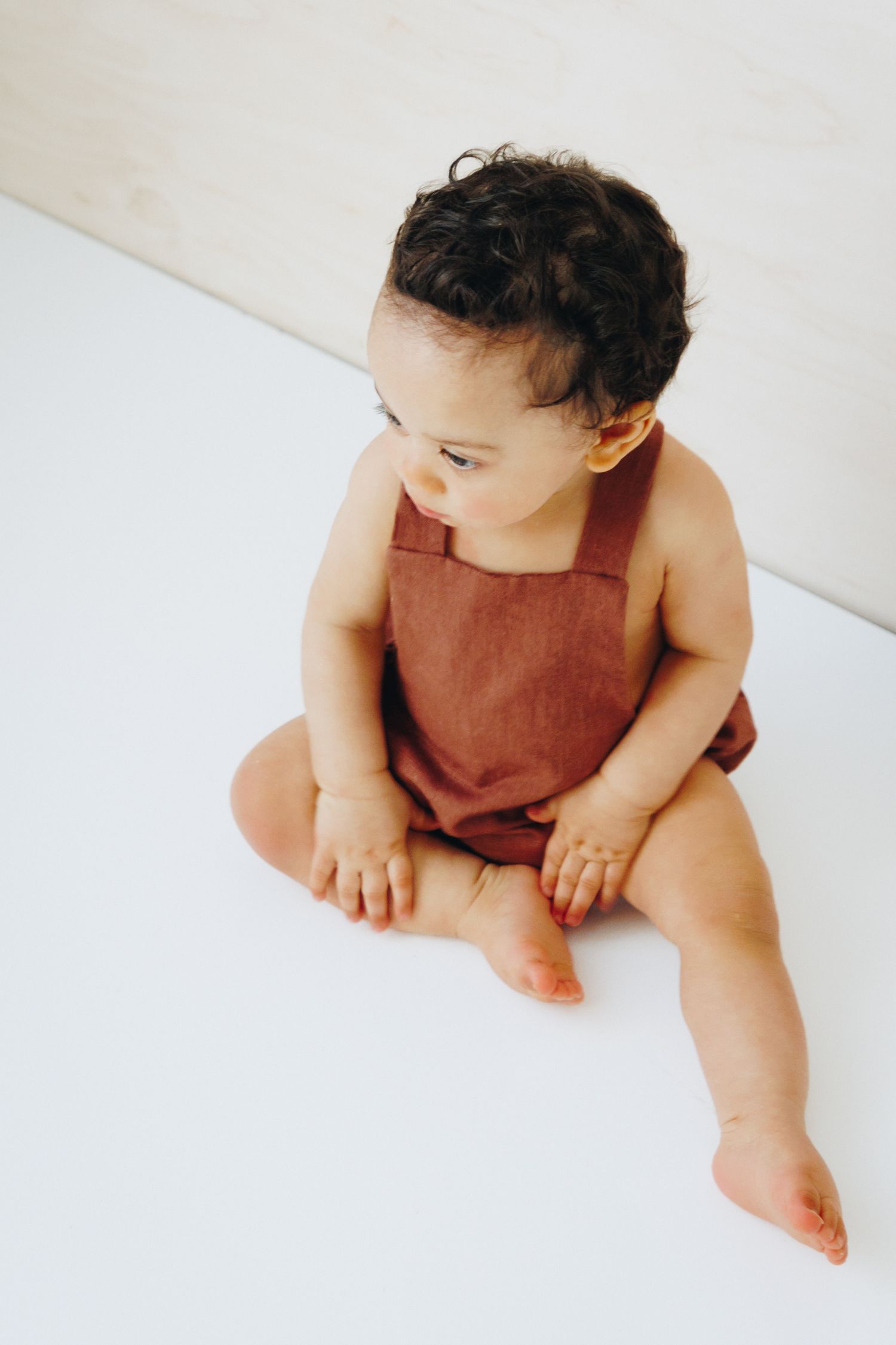 Baby sitting on white floor wearing a rust brown linen romper, looking to the side.