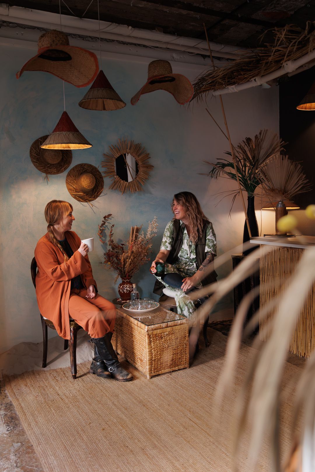 Two women sit in a bohemian-styled room with wicker decor, hats on wall, chatting over drinks.