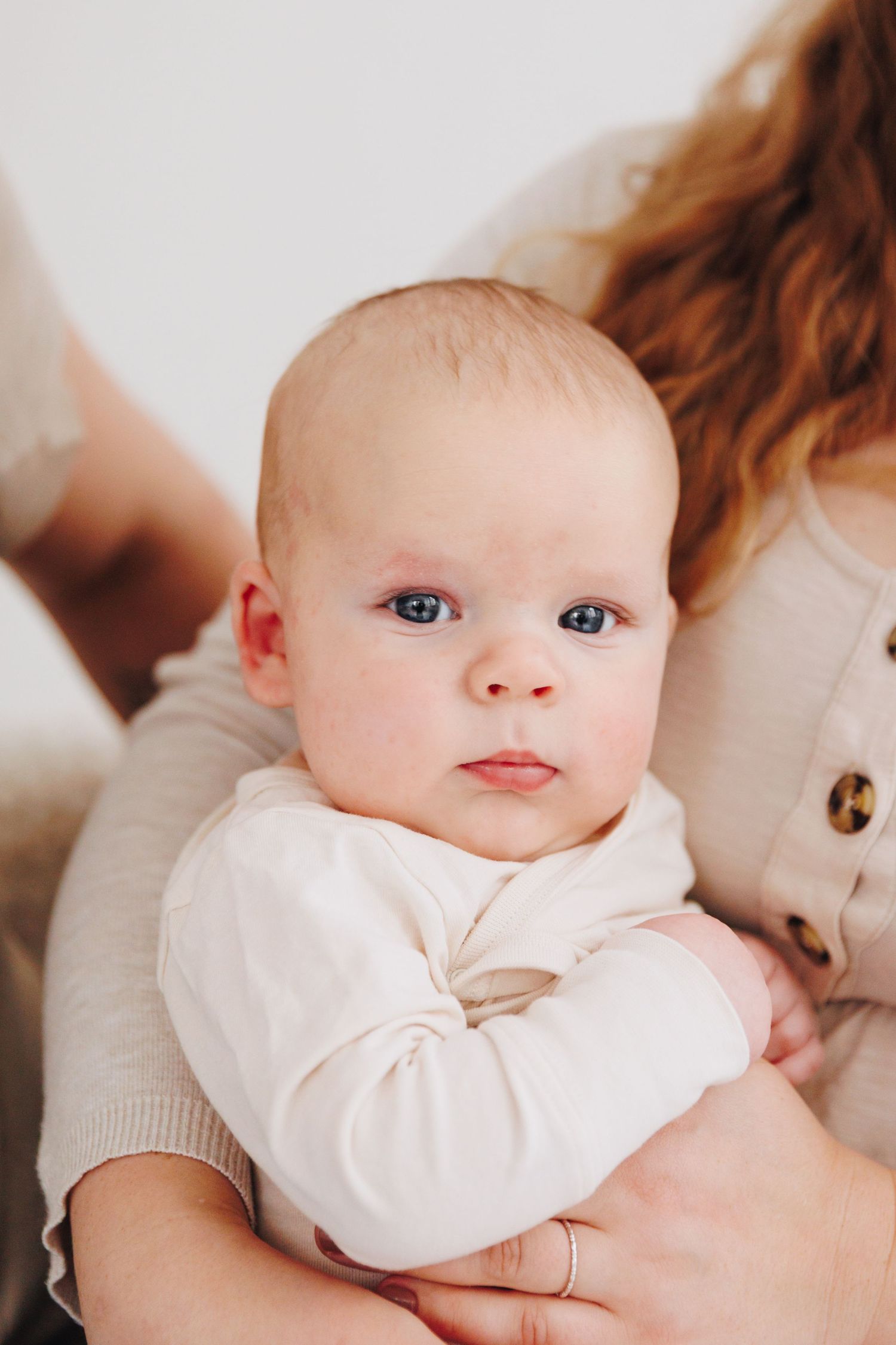 A newborn baby with blue eyes in a white onesie being cradled by a redheaded mother wearing a beige outfit.