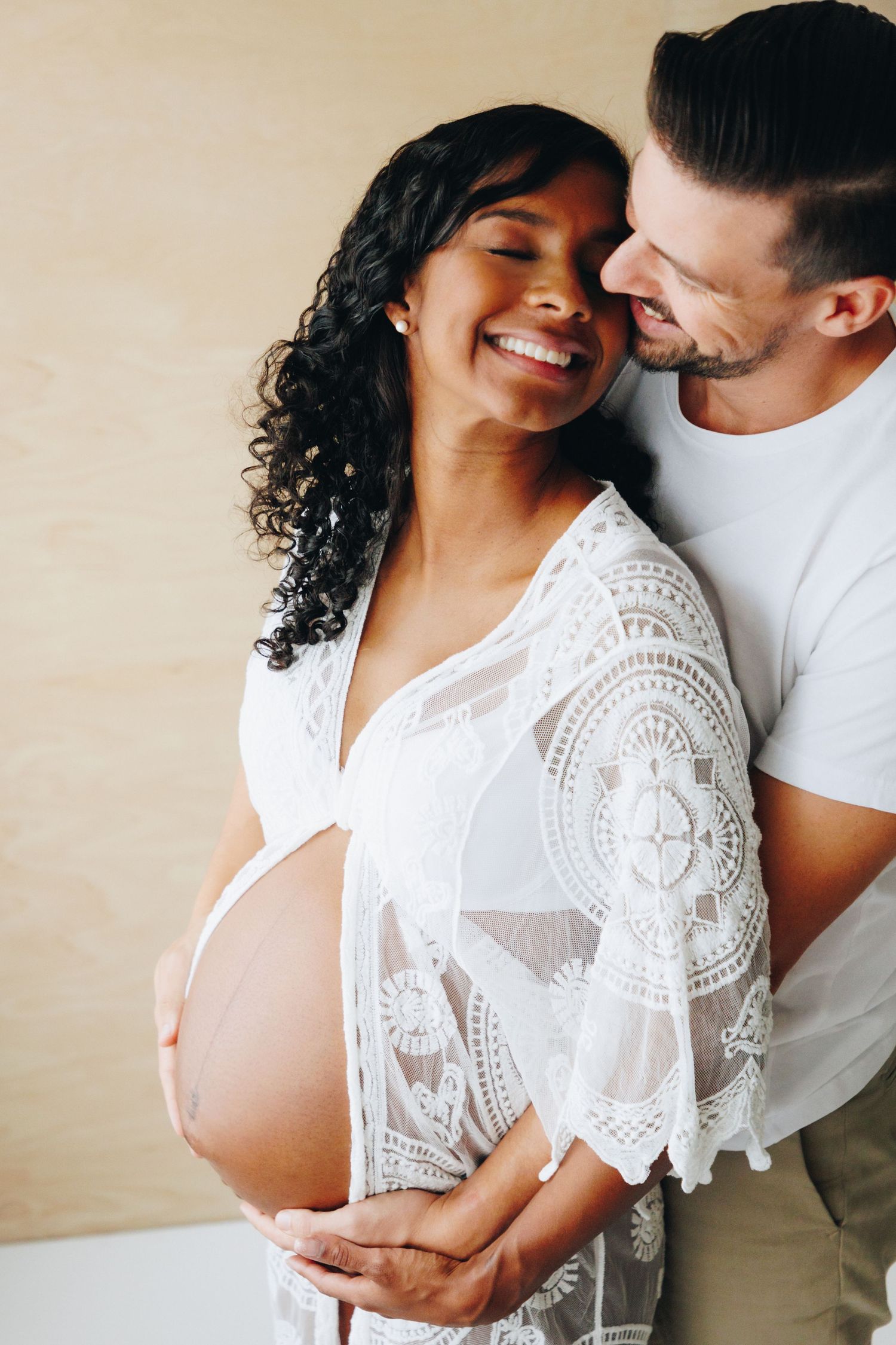 A romantic couple embraces in intimate maternity photos wearing white against a neutral background.