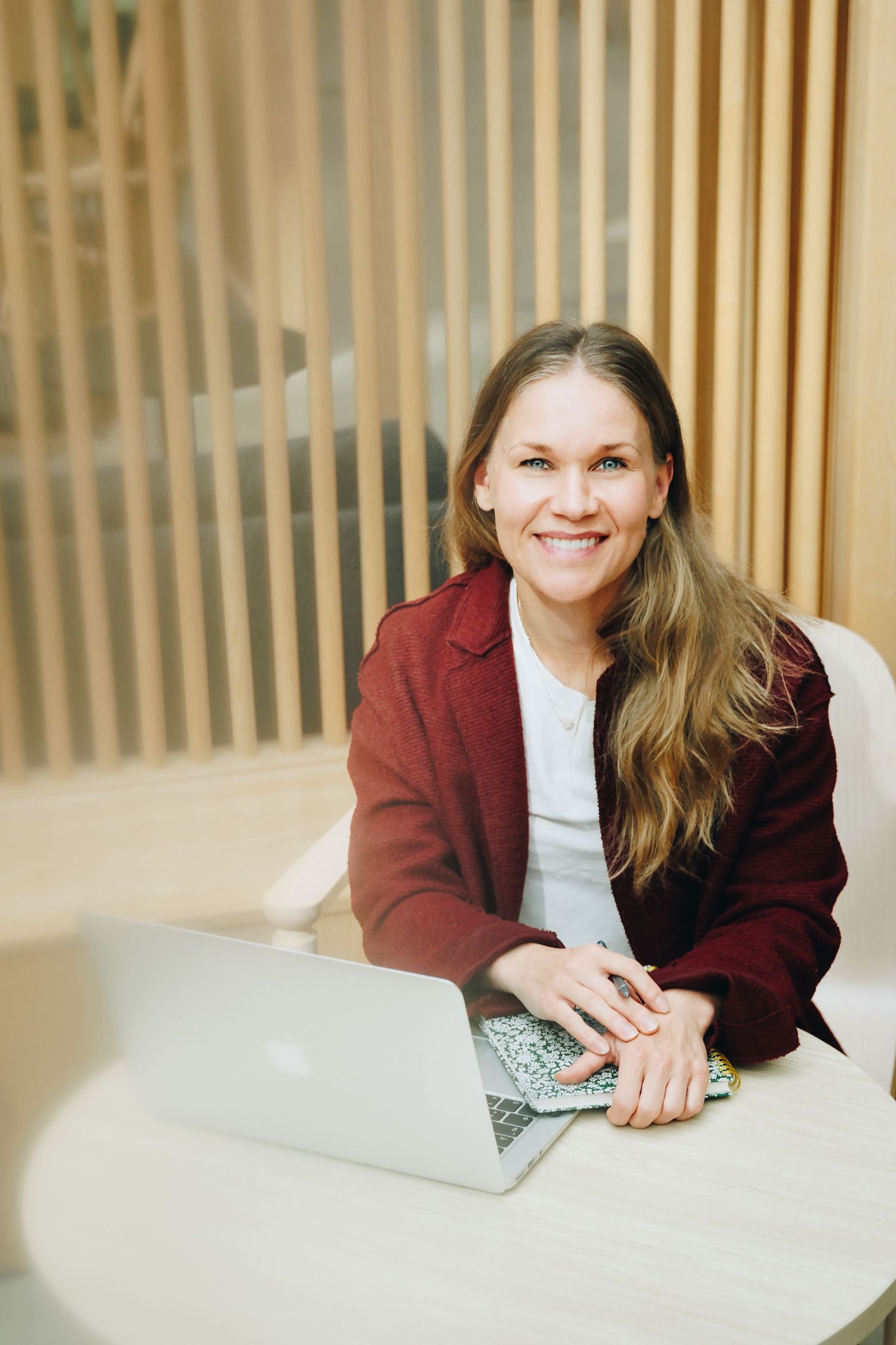 Professional wearing a burgundy blazer and white top sits at desk with laptop in bright office setting.