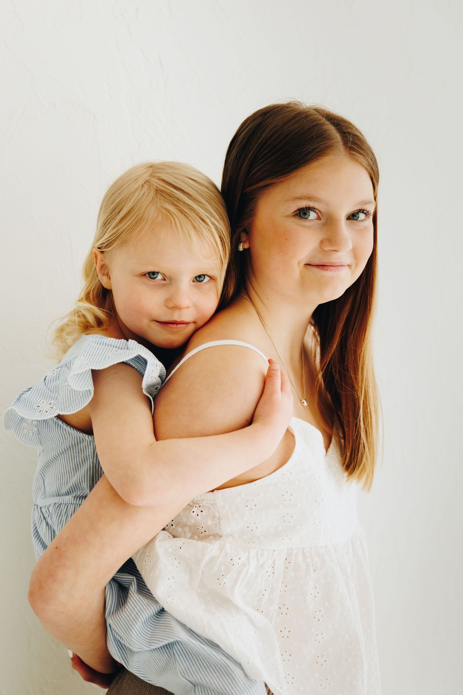 Older girl with brown hair in white dress giving piggyback ride to young blonde toddler in blue dress against white background.