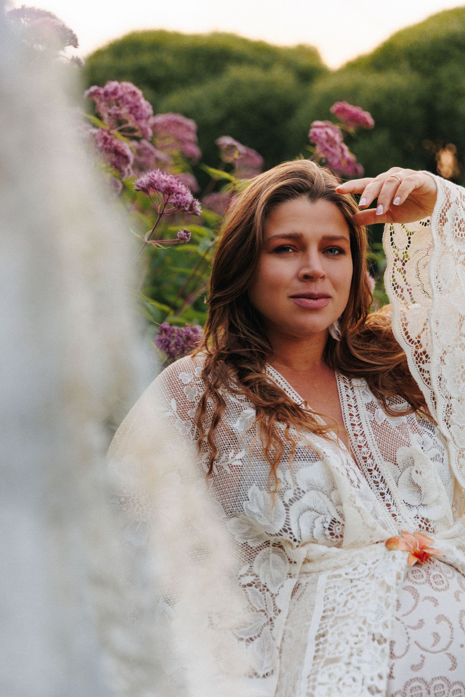 Woman in white lace dress poses among purple blooming flowers in a garden at golden hour.