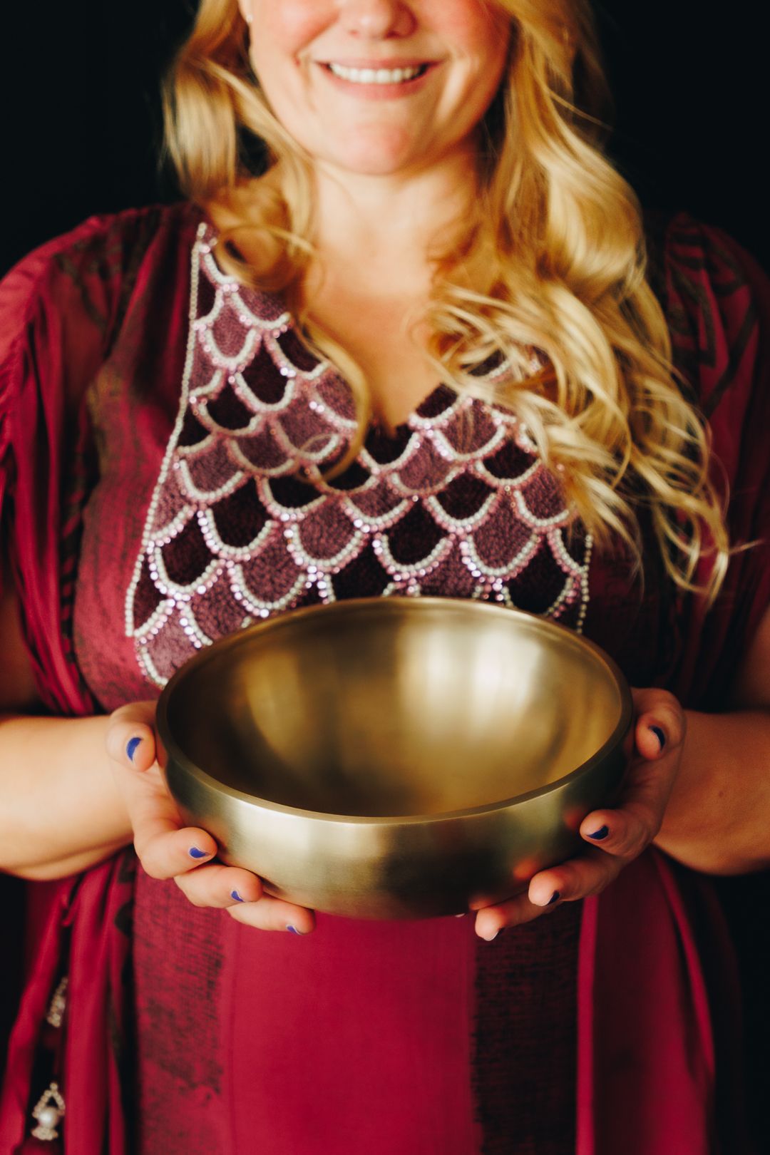 Smiling blonde woman in red dress holding a golden singing bowl against dark background.