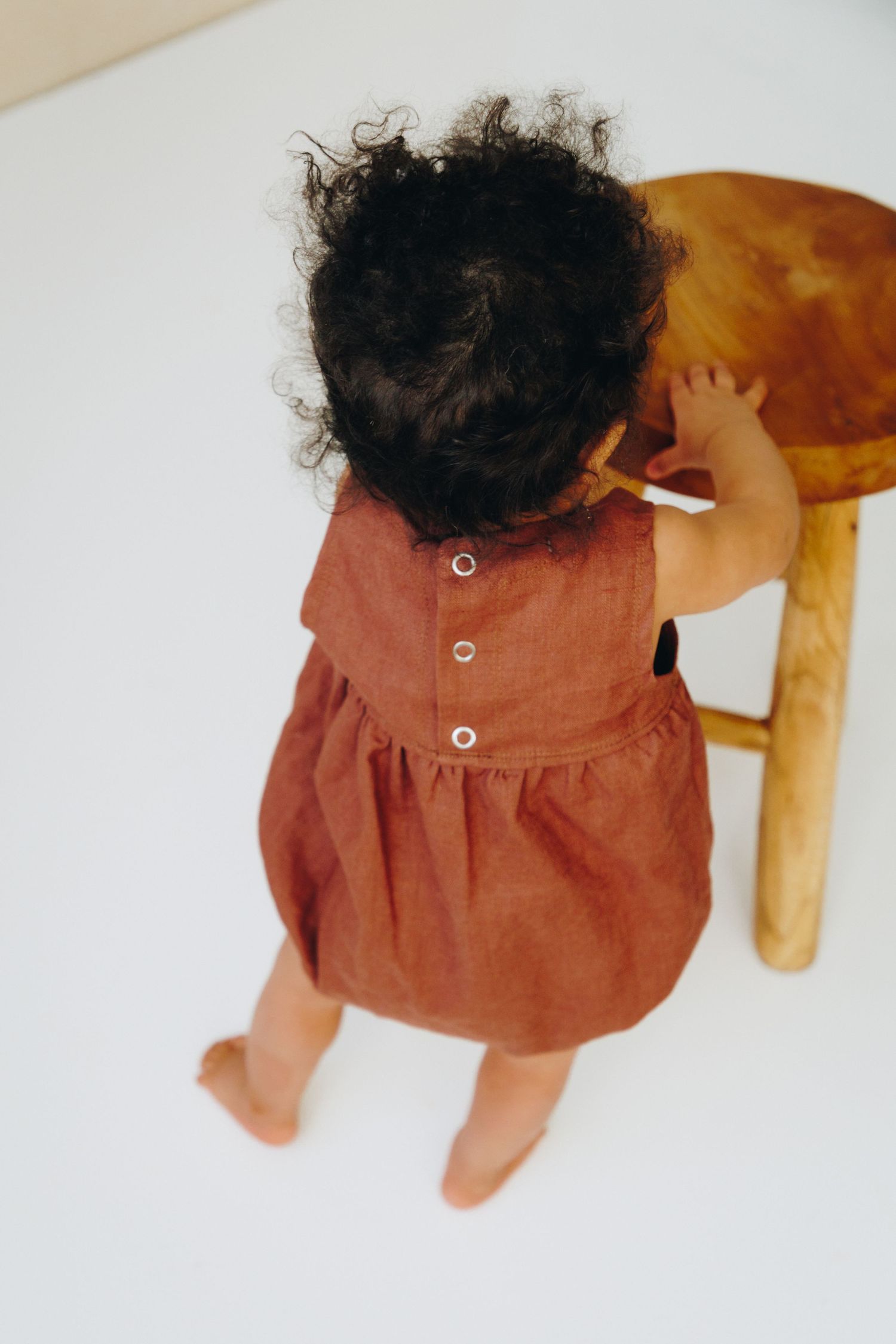 Toddler girl in rust linen dress standing barefoot, viewed from above, reaching toward a wooden stool.
