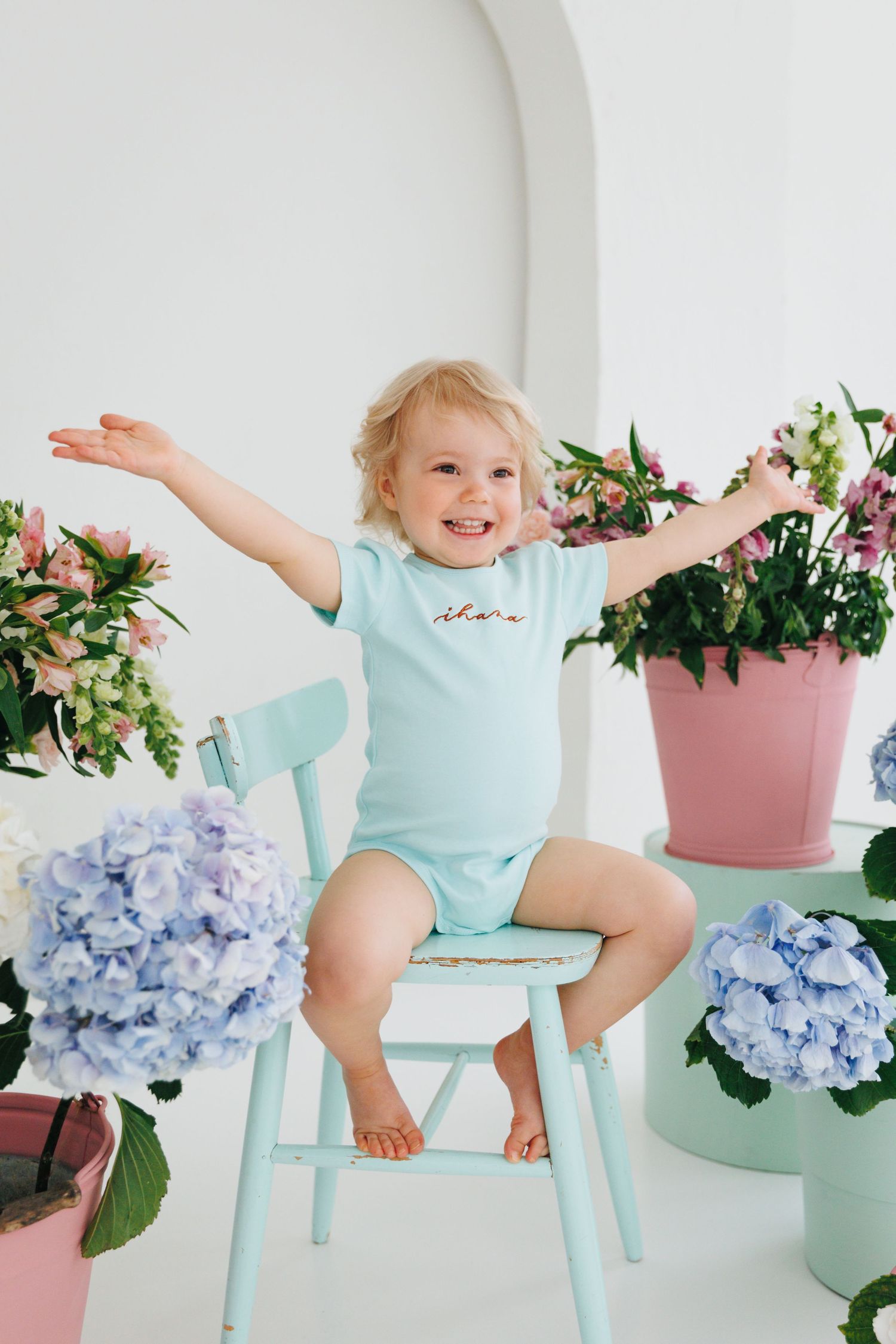 Happy toddler in mint bodysuit sits on teal chair surrounded by colorful flowers including hydrangeas.