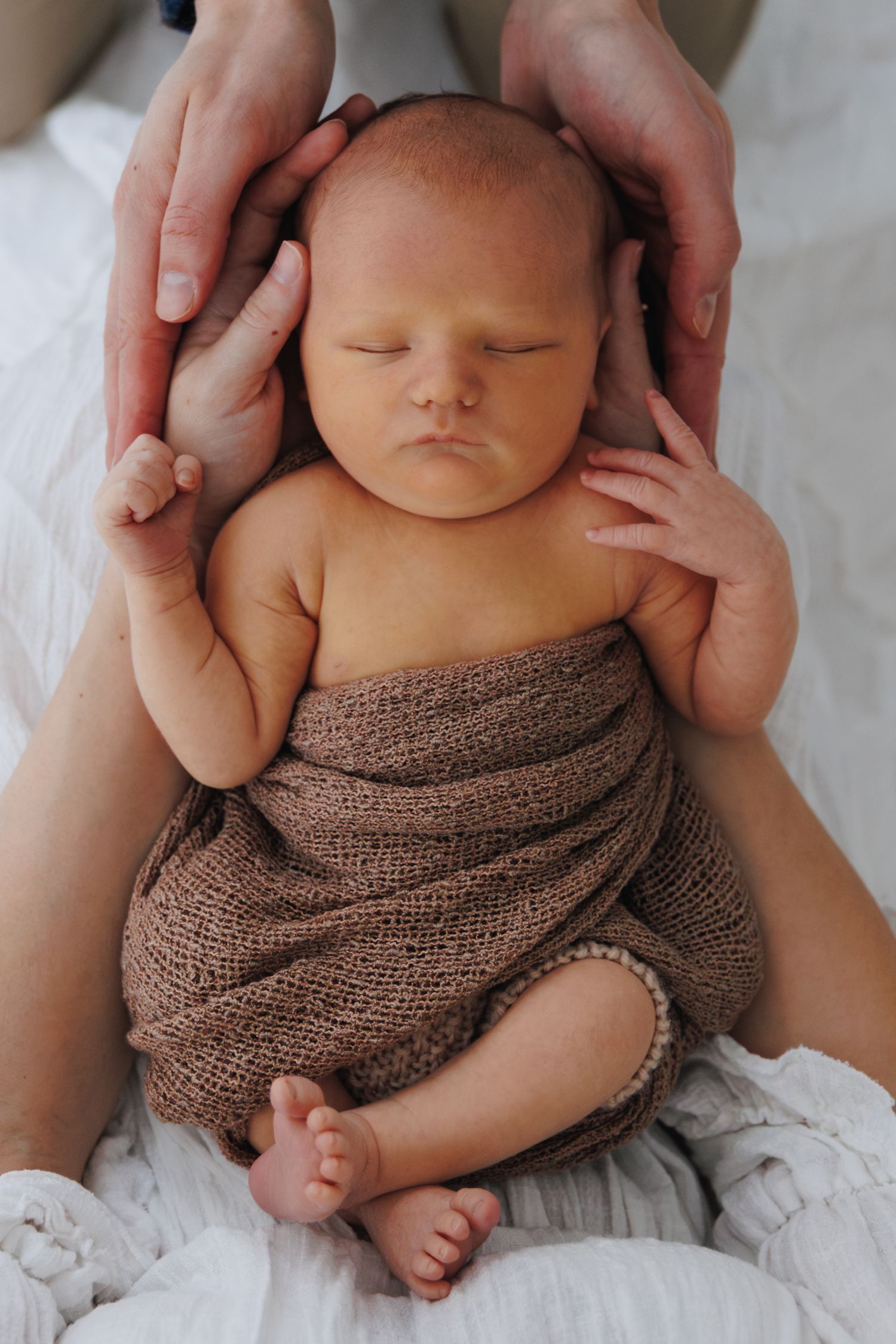 Newborn baby wrapped in brown knit swaddled peacefully while parent hands gently cradle the infant's head.