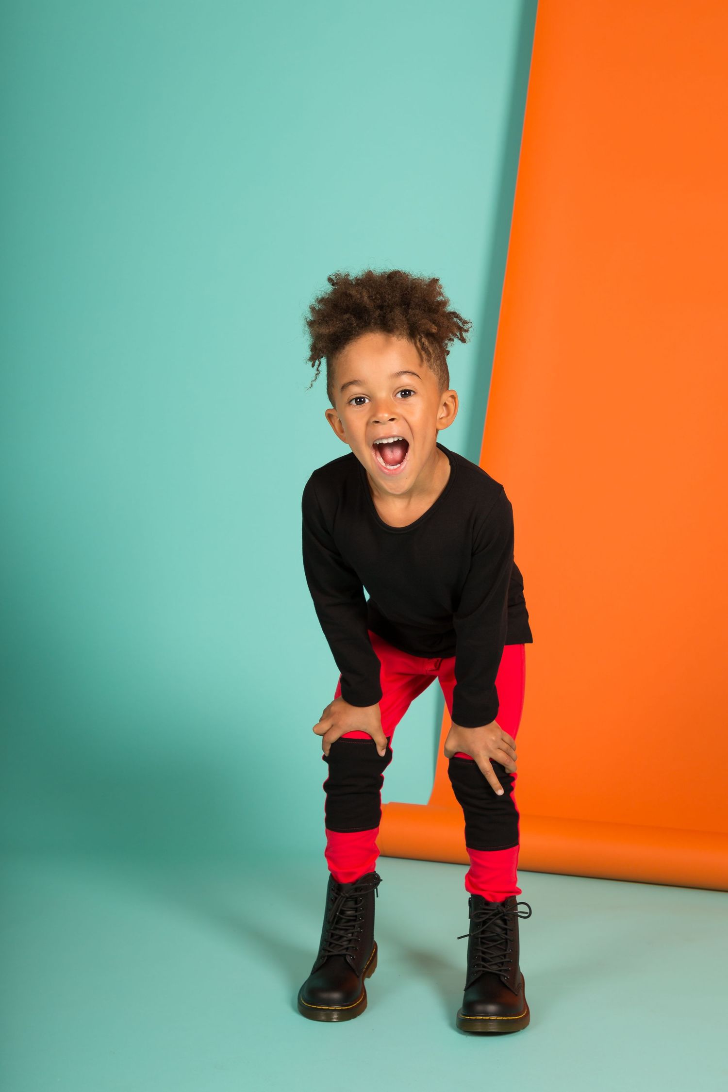 Energetic child wearing black top and red pants strikes playful pose against turquoise and orange background.