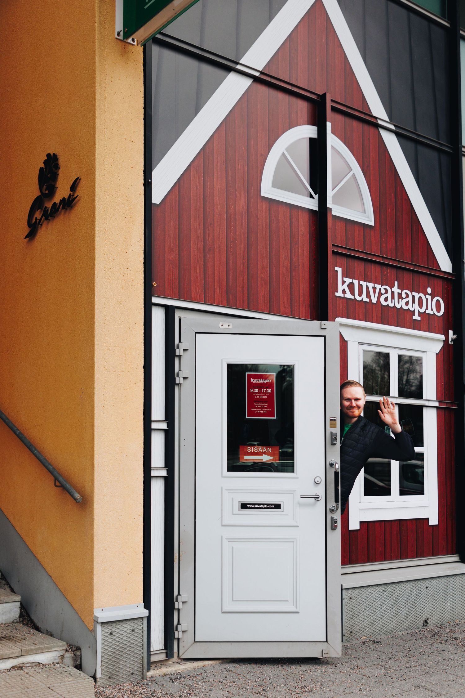 Man waving from doorway of red barn-style Kuvatapio building with white trim in Finland.