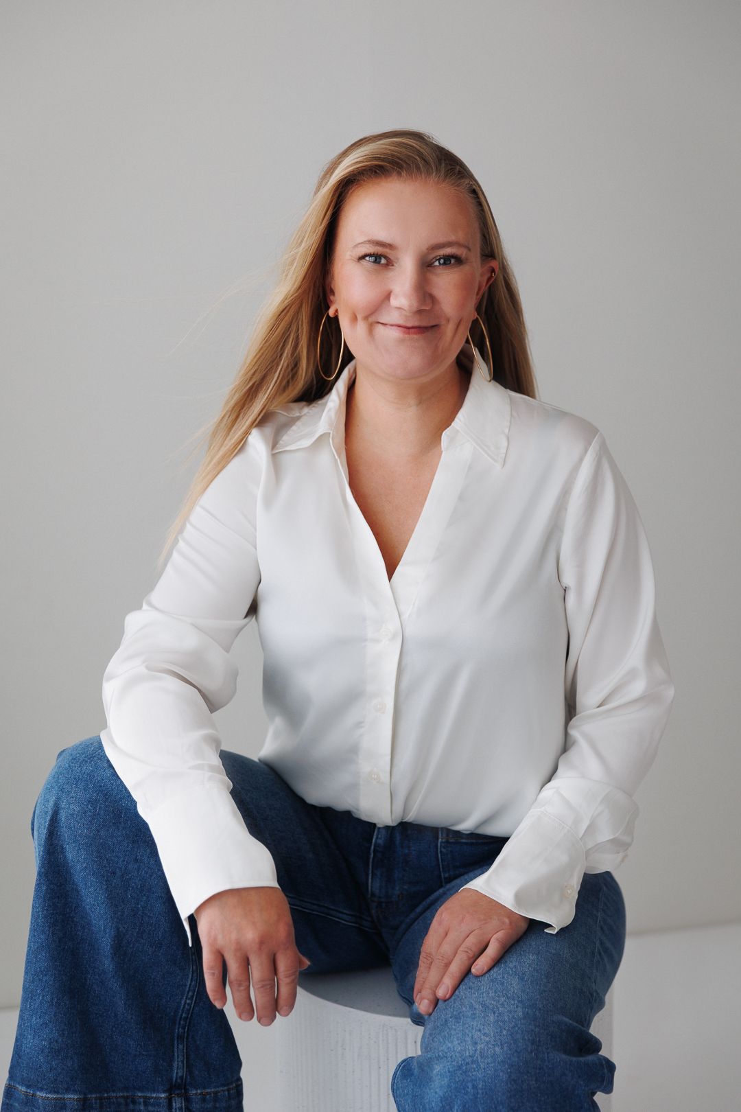 Smiling woman in white blouse and jeans sitting relaxed against a neutral grey background.