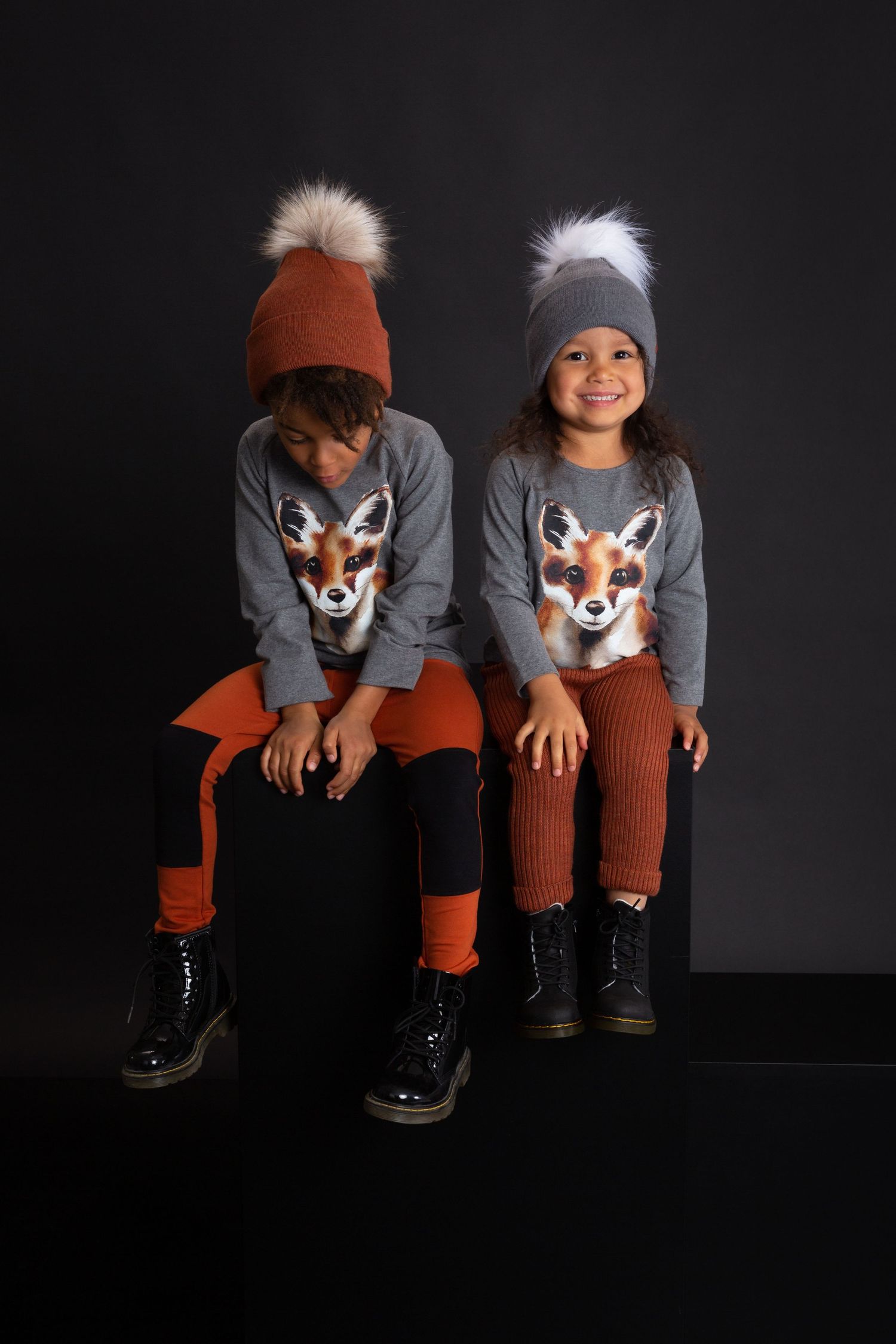 Two kids in matching gray fox sweaters, orange leggings, and pom-pom beanies sit together against a dark background.