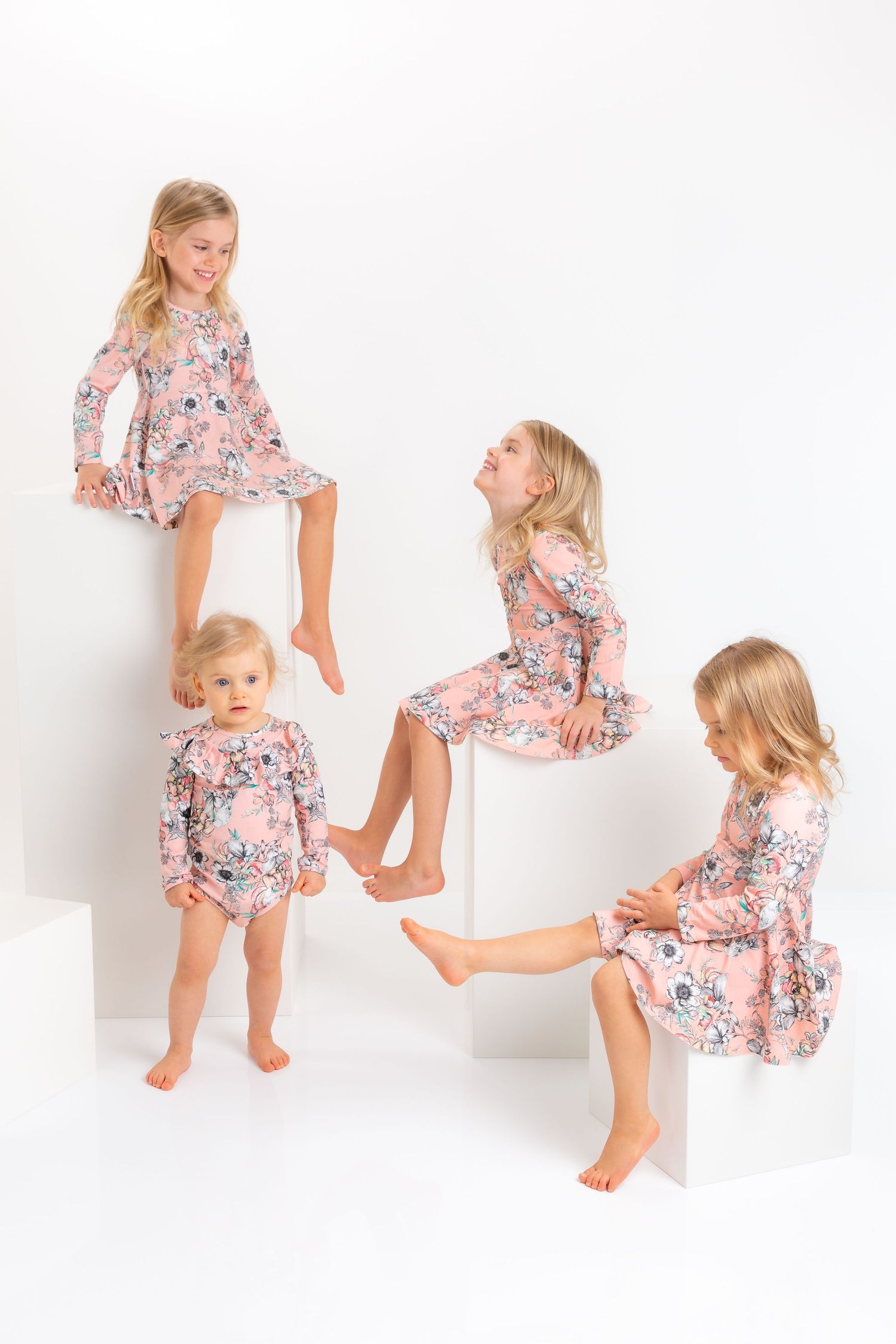 Four young girls in matching pink floral dresses and romper posed on white geometric blocks in a studio.