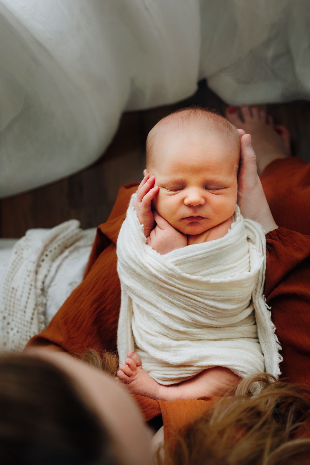 A sleeping newborn swaddled in white fabric is photographed in a warm and intimate setting.