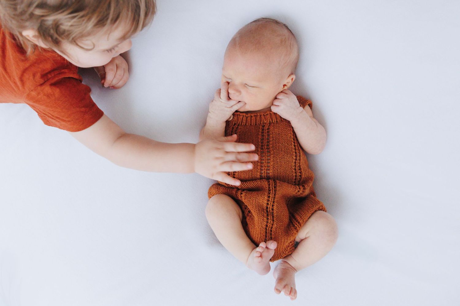 A toddler in a red shirt gently touches a sleeping newborn baby wearing a knit rust-colored romper on a white surface.
