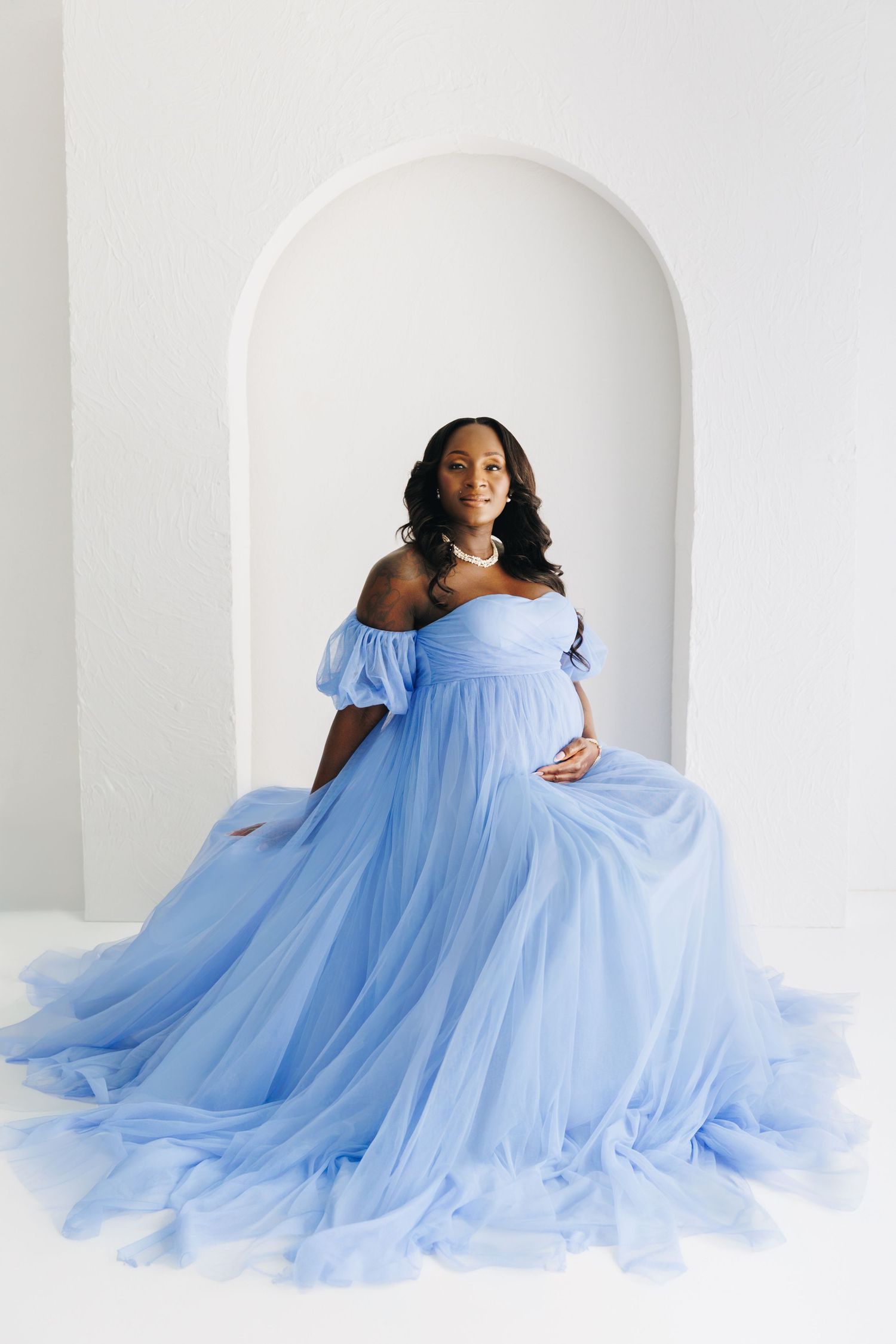 Pregnant woman in flowing off-shoulder blue tulle gown poses gracefully in white arched studio setting.