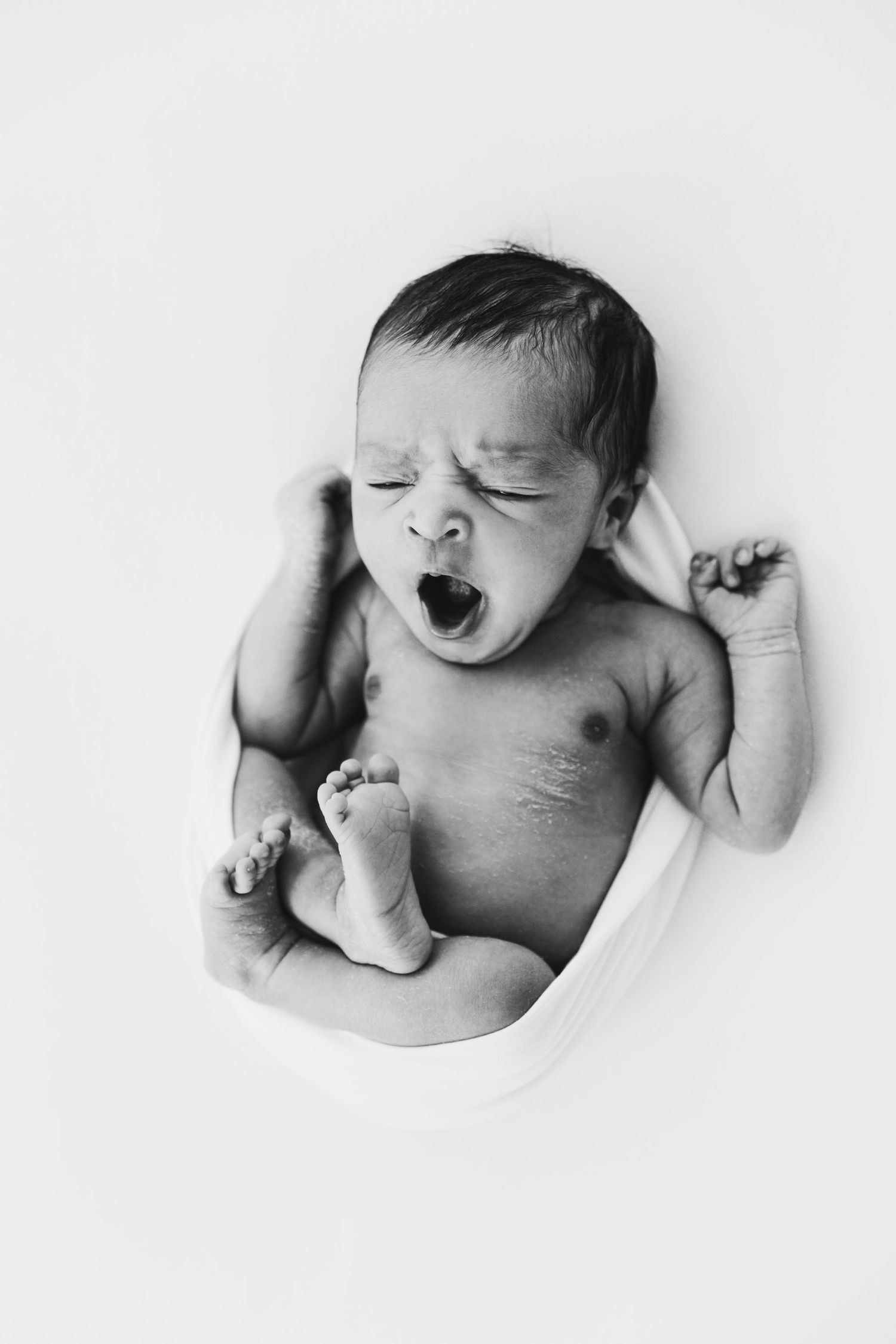 Black and white photo of a newborn baby yawning and stretching on a white blanket.