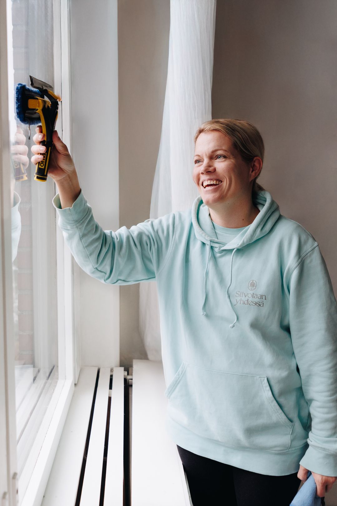 Smiling woman in mint hoodie cleaning window with electric brush tool, indoor home cleaning service.