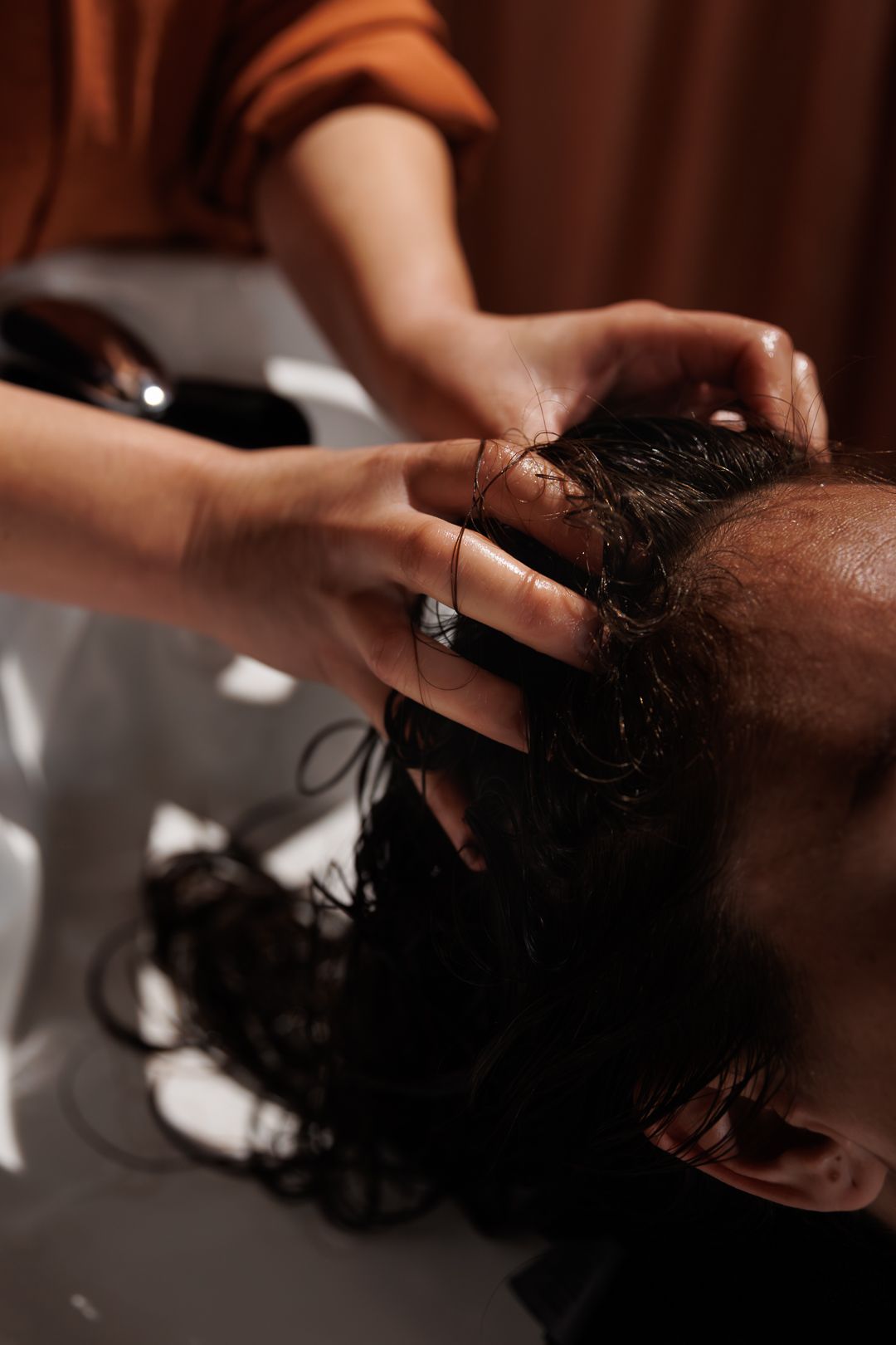 A hairstylist massages shampoo into a client's dark wet hair at a salon washing basin.