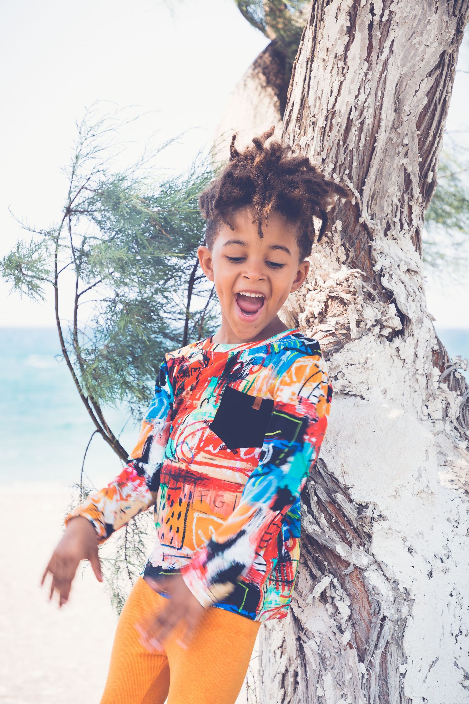 Joyful child in colorful top and orange pants laughing beside a tree near the beach.