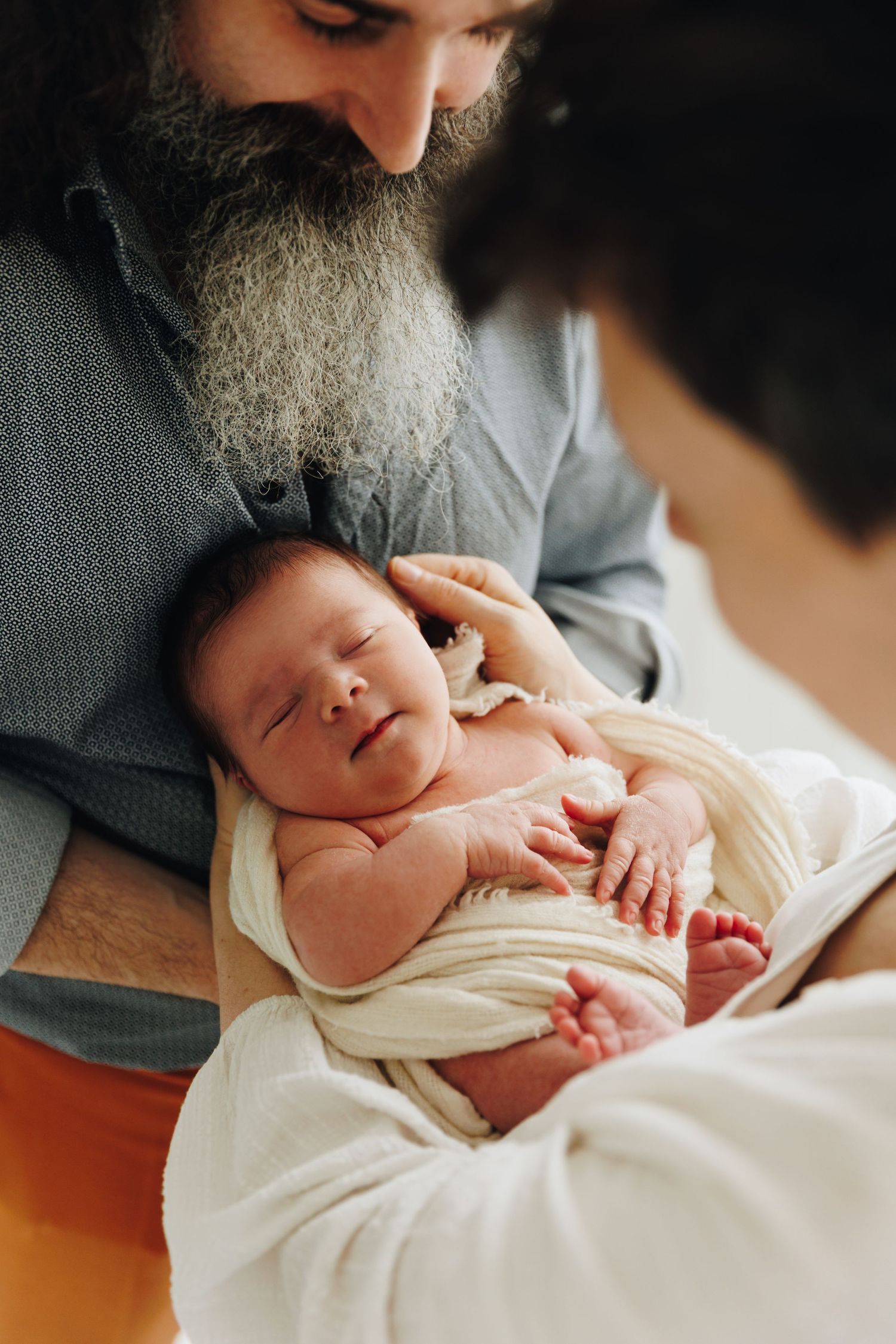 Bearded grandfather smiling at sleeping newborn baby wrapped in white blanket held by parent. Vastasyntyneen kuvaus Helsingissä.