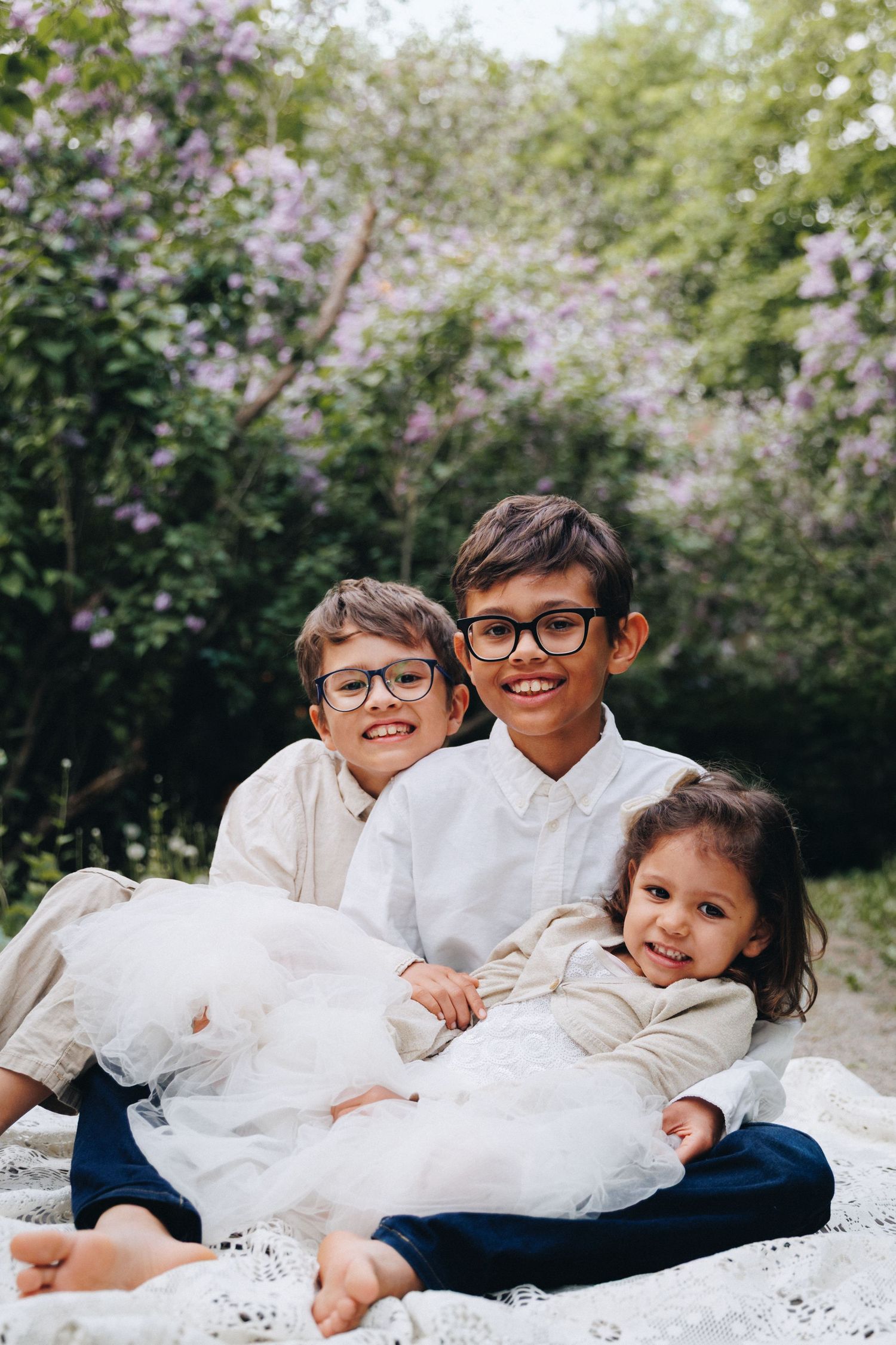 Three smiling children sitting on a blanket outdoors near blooming lilac bushes, two boys wearing glasses and a little girl.