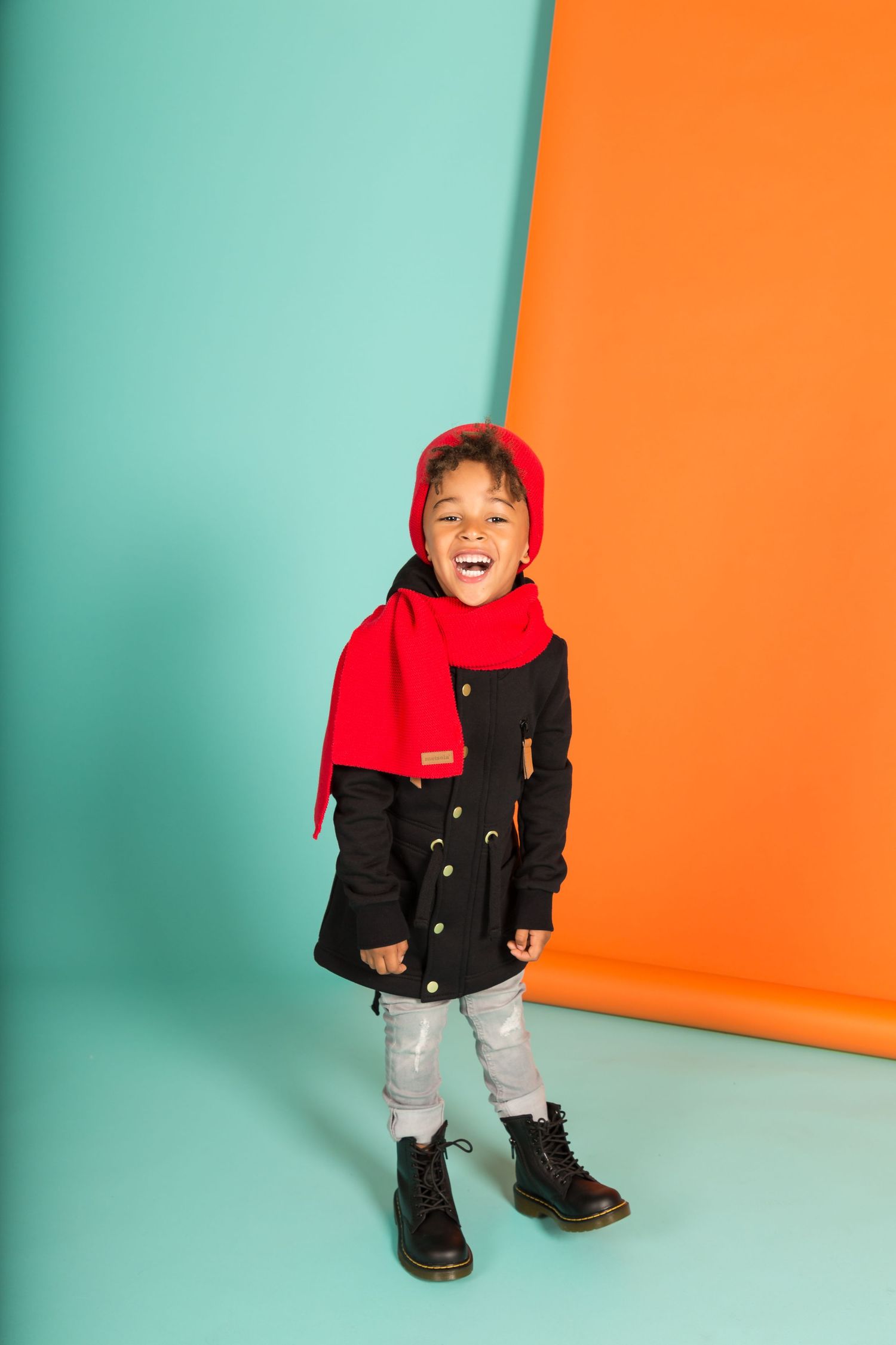Smiling child in black coat, red scarf and hat, gray pants, black boots, posing against teal and orange background.