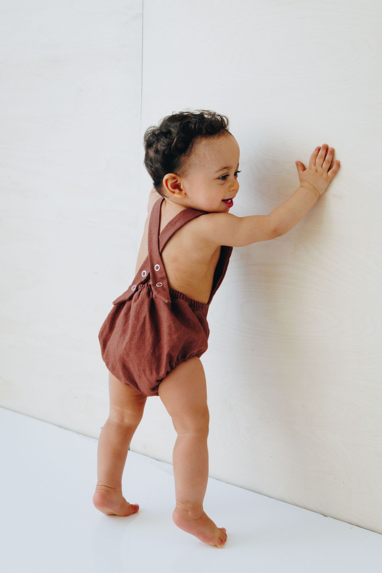 Toddler in brown halter romper standing against white wall, reaching out with one hand.