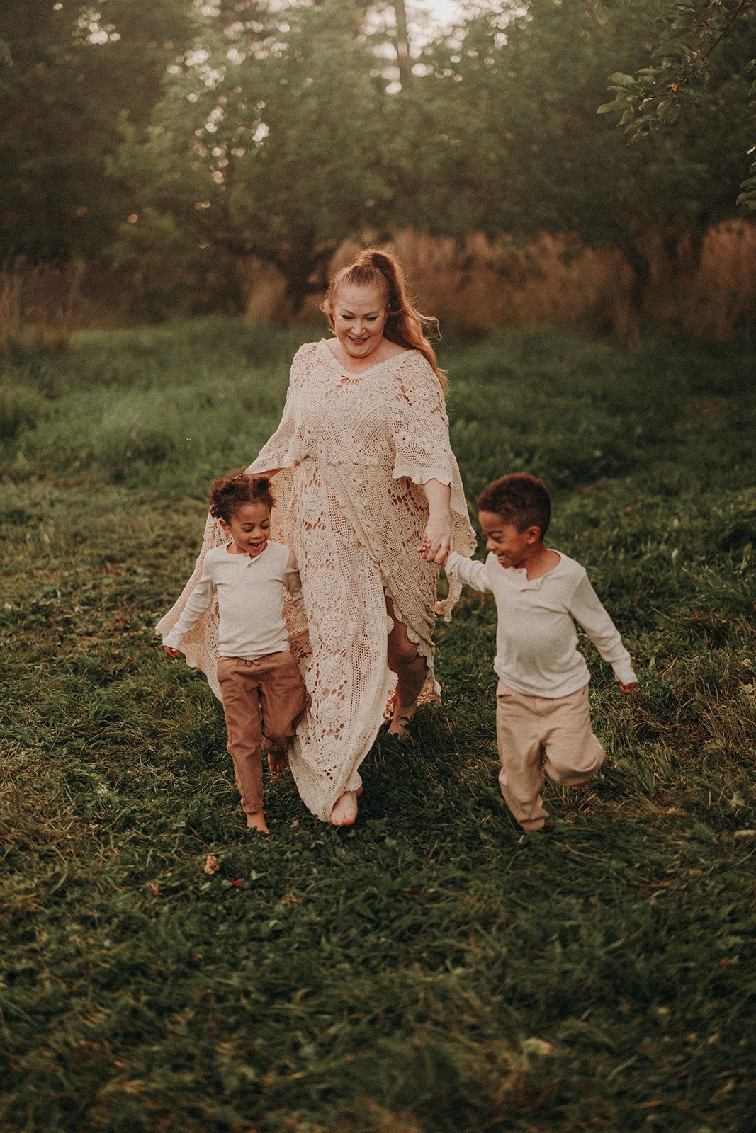 A family in white lace and cream outfits runs playfully through a grassy field at sunset.
