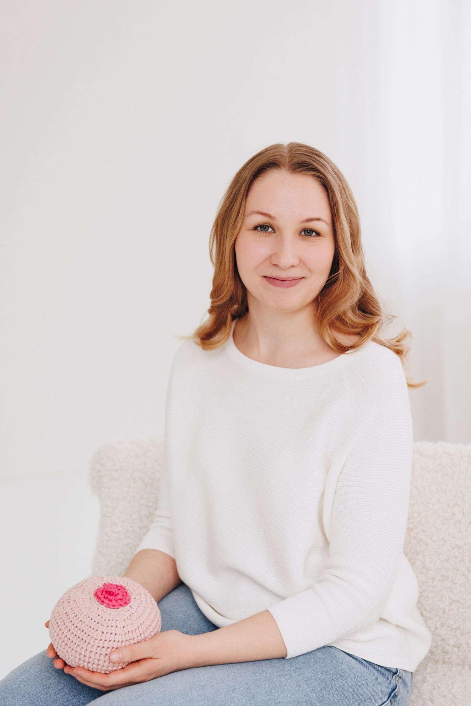 Woman in white sweater sitting on chair holding a pink crocheted breast model for breastfeeding education.