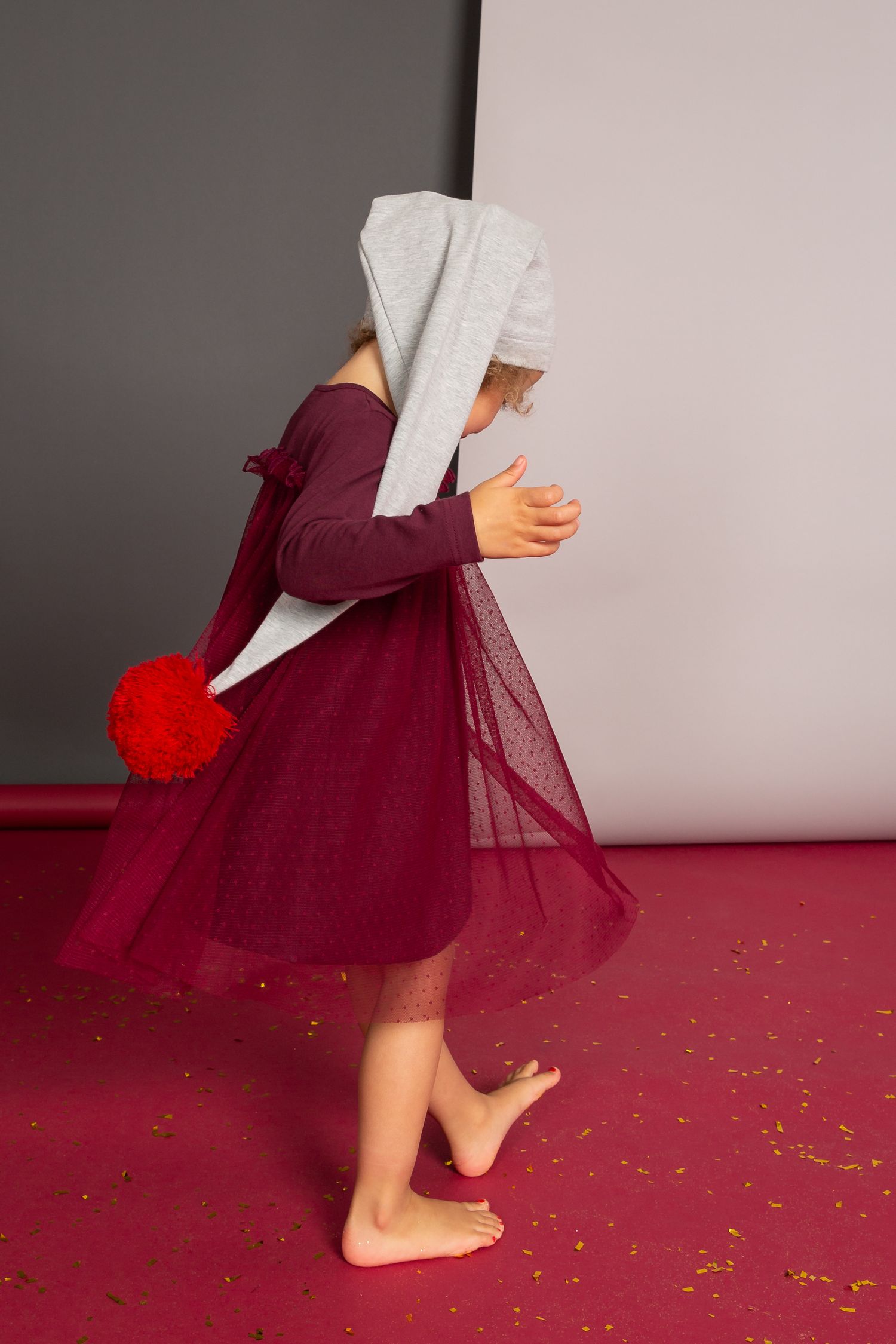 Young girl in burgundy tulle dress and gray bunny hat holds red pom pom, dancing barefoot on red floor.