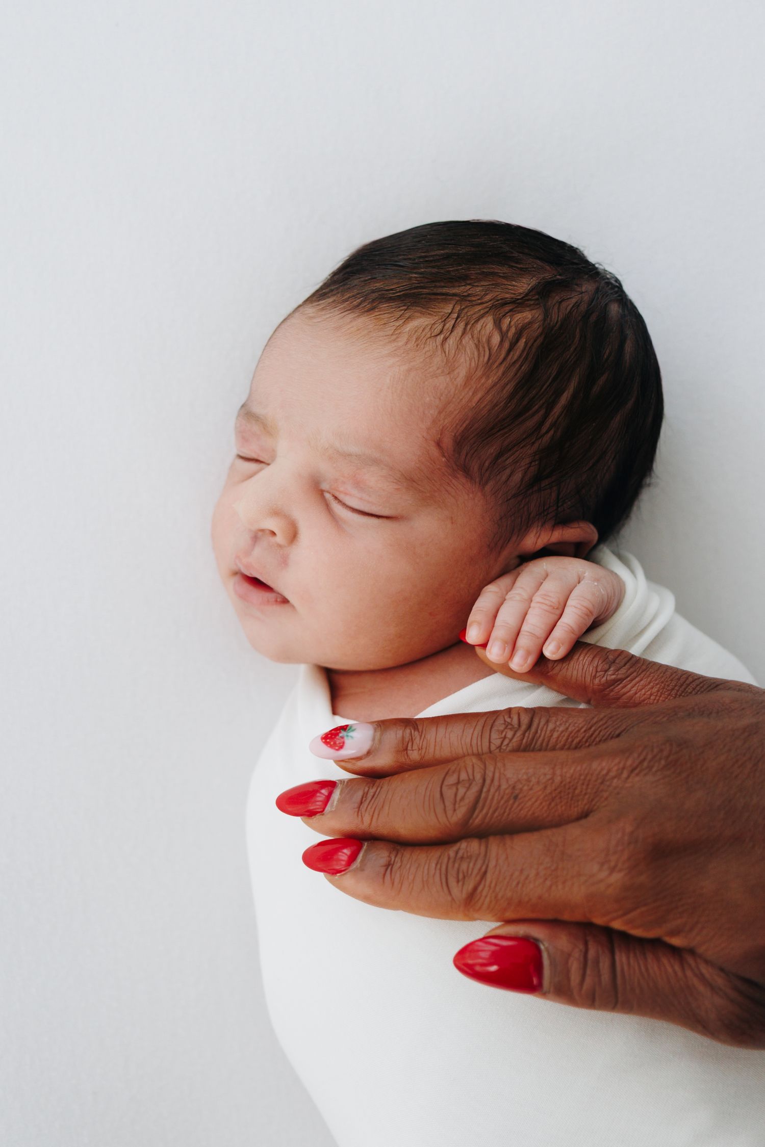 Newborn baby sleeping peacefully while wrapped in white, with a hand featuring red nails gently cradling the infant.
